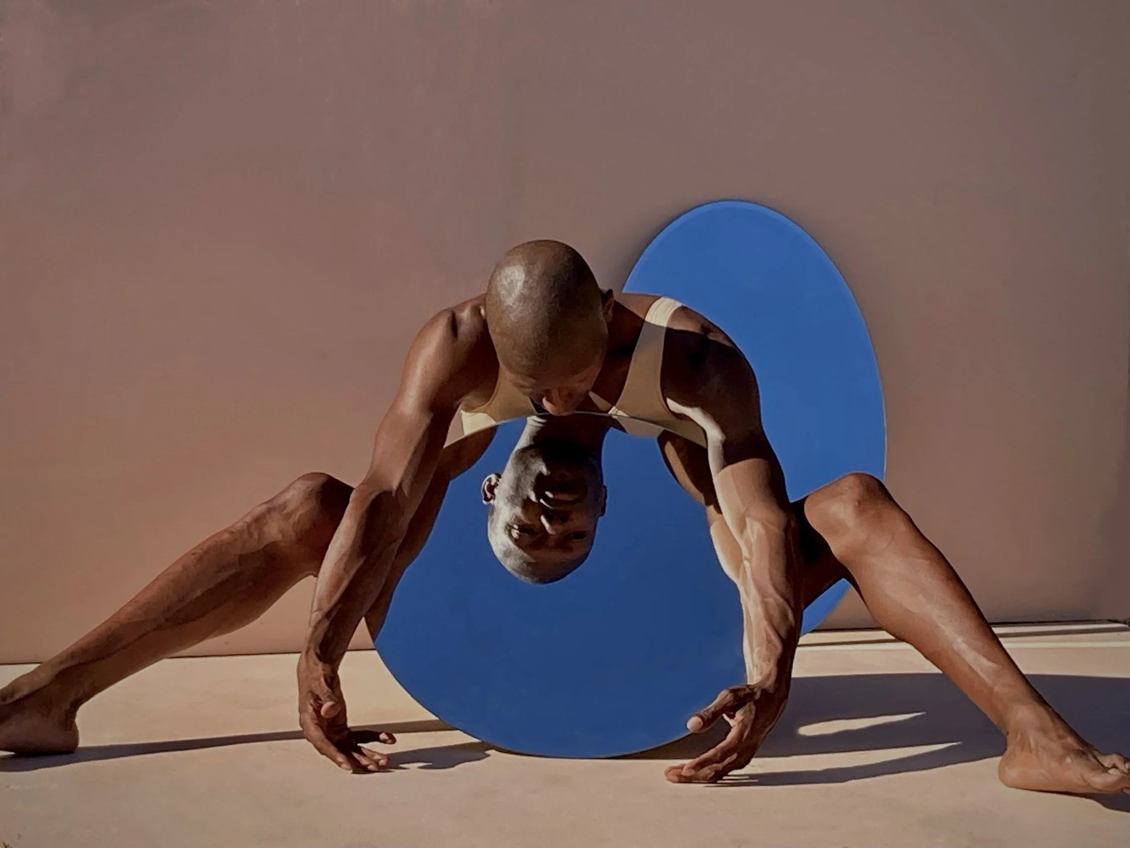A person with a shaved head and dark skin performing a pose on the floor, with legs spread wide and hands touching the ground, positioned behind a large mirror.