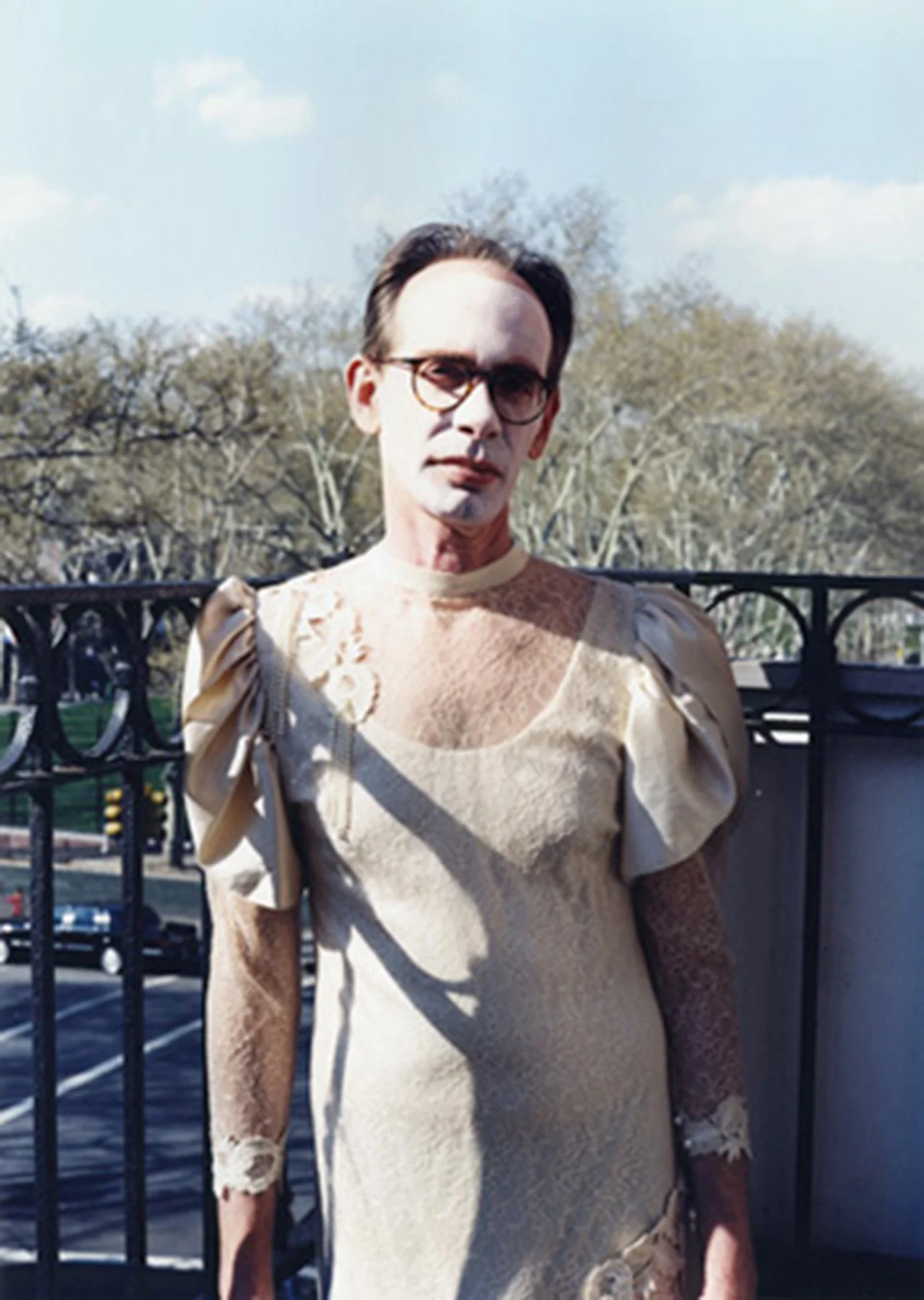 A man with dark hair, glasses, and pale skin standing outdoors next to a black metal fence, with trees and a parking lot in the background, wearing a worn, vintage ivory dress with puffy sleeves.