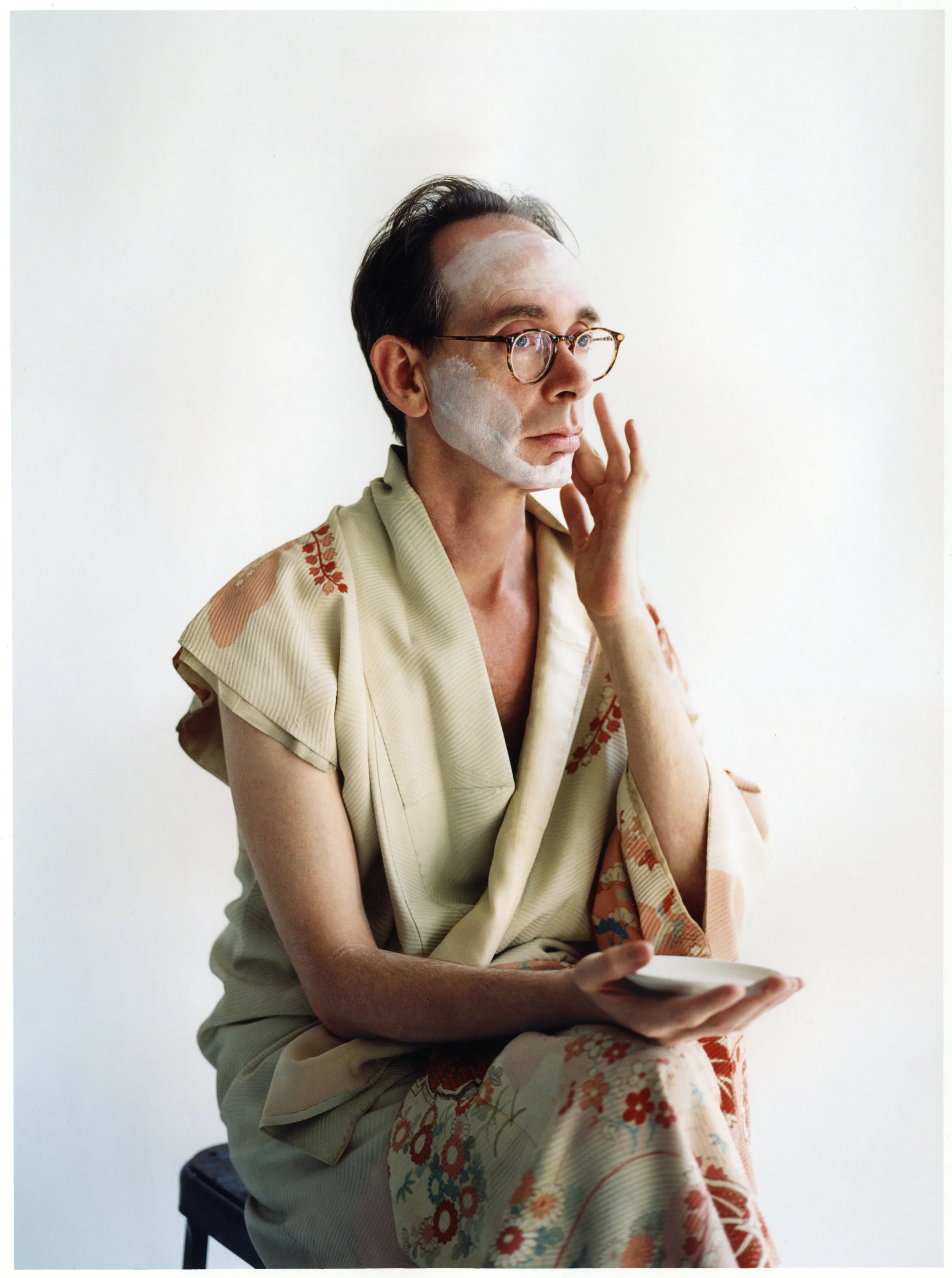 Musicuian Arto Lindsay with glasses sitting on a stool, applying white face makeup and holding a makeup palette, dressed in a kimono with floral patterns, against a plain white background.