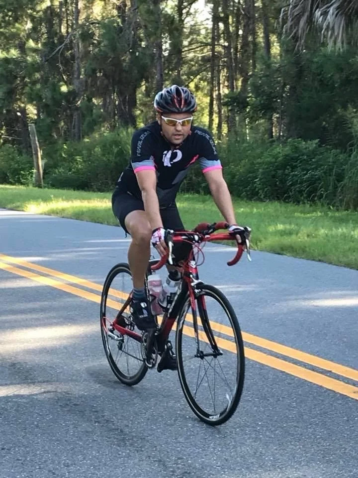 Man riding a road bike on a paved country road surrounded by trees, wearing a black helmet, cycling sunglasses, and a black cycling outfit with pink accents.