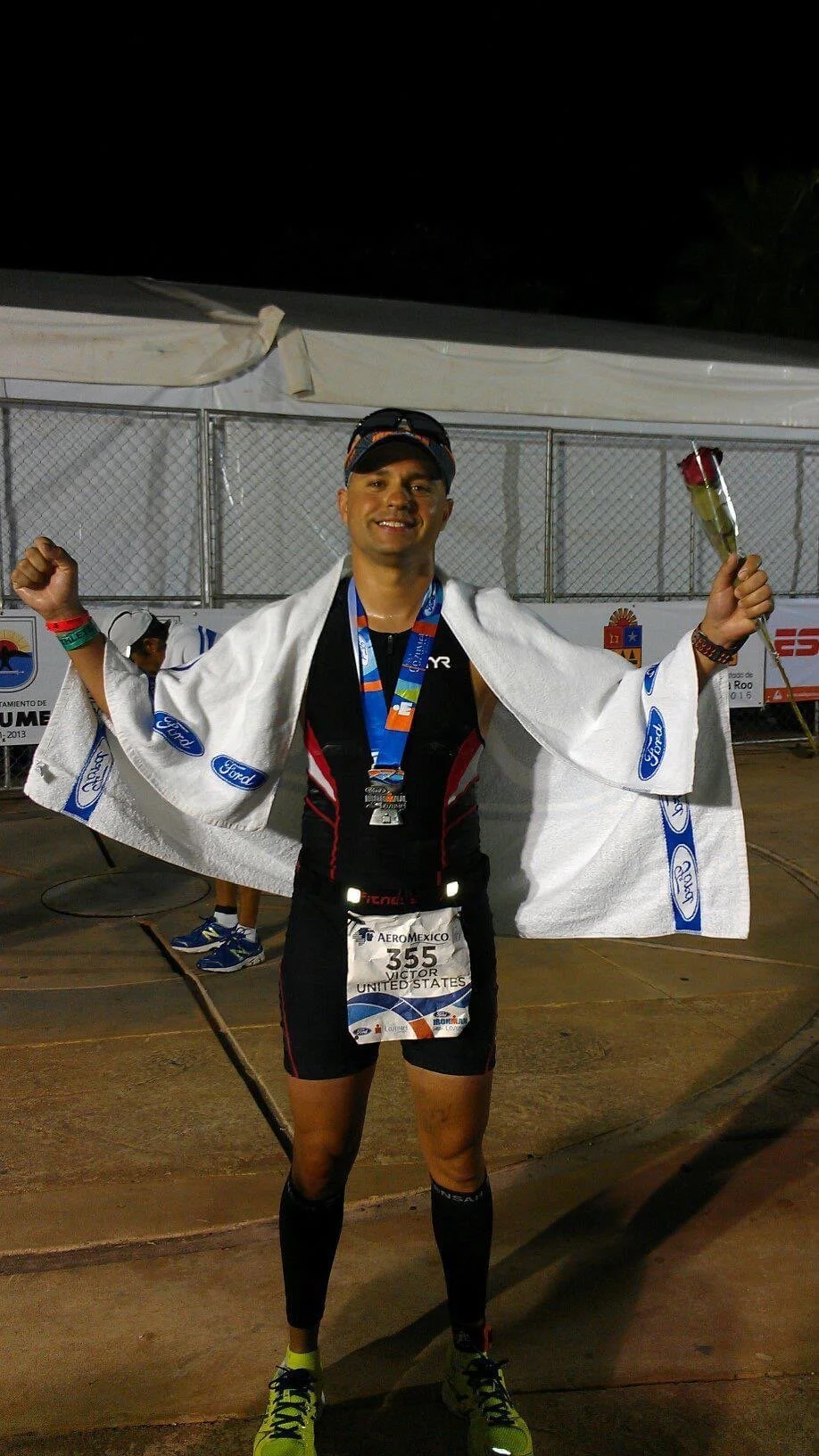 A male marathon runner celebrating at the finish line, holding a medal and a rose, wearing a race bib that reads 'Victor United States,' with a white towel over his shoulders during nighttime.