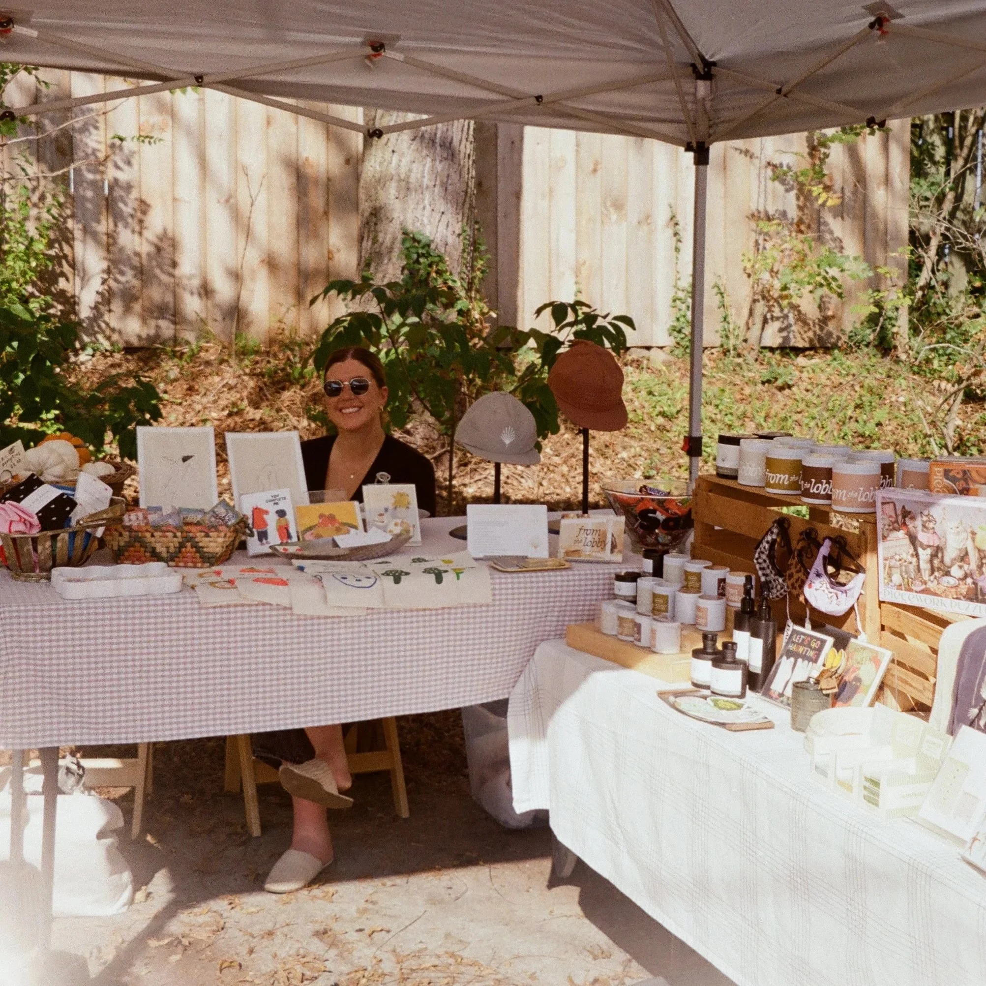 A woman sits at a craft vendor booth outdoors, wearing sunglasses and smiling, with handmade items and artwork on display, shaded by a canopy.