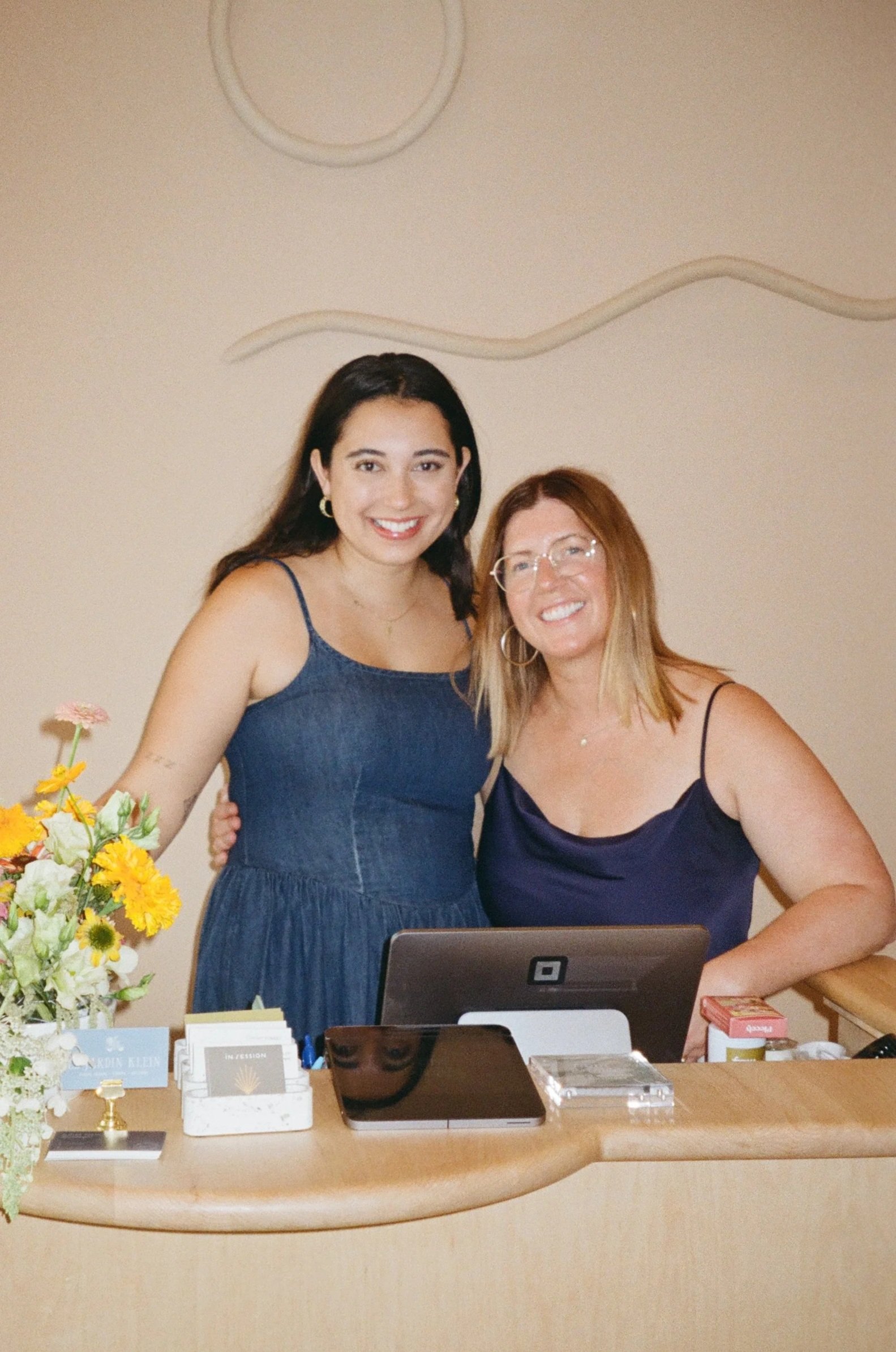 Two women smiling at a reception desk surrounded by flowers and office supplies.