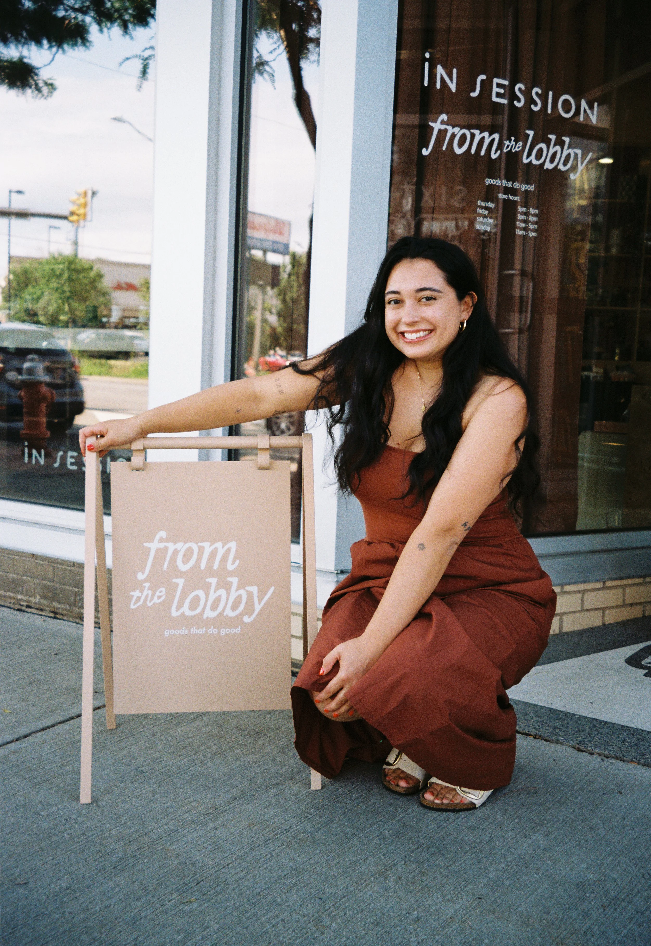 A woman with long black hair, wearing a rust-colored dress and sandals, crouching next to a beige sign that reads 'from the lobby' outside a store, smiling at the camera.