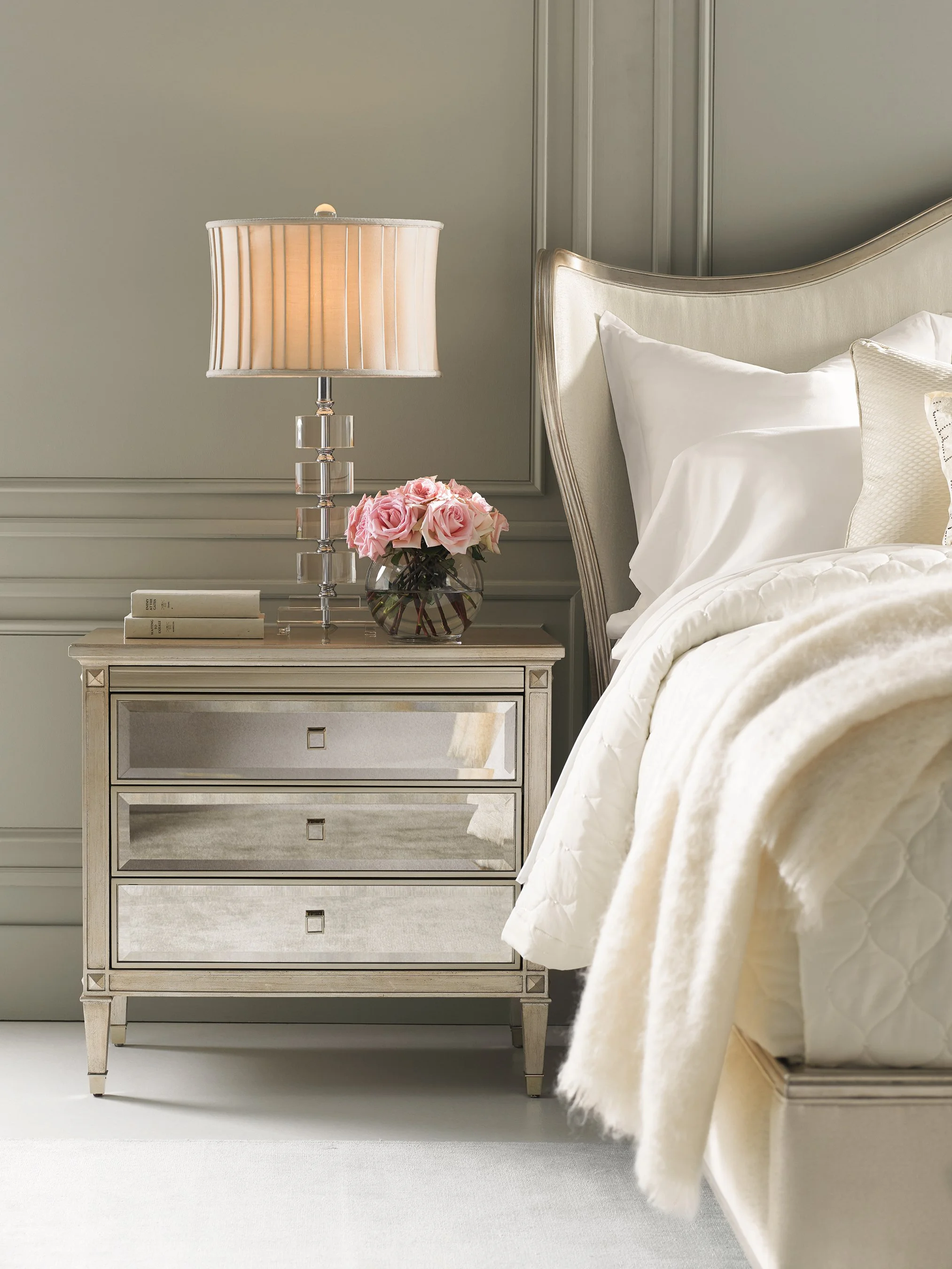 Hollywood Glam bedroom vignette featuring a mirrored three-drawer nightstand, crystal-accent table lamp, curved upholstered headboard, soft neutral bedding, and fresh pink roses, creating an elegant and sophisticated luxury aesthetic.