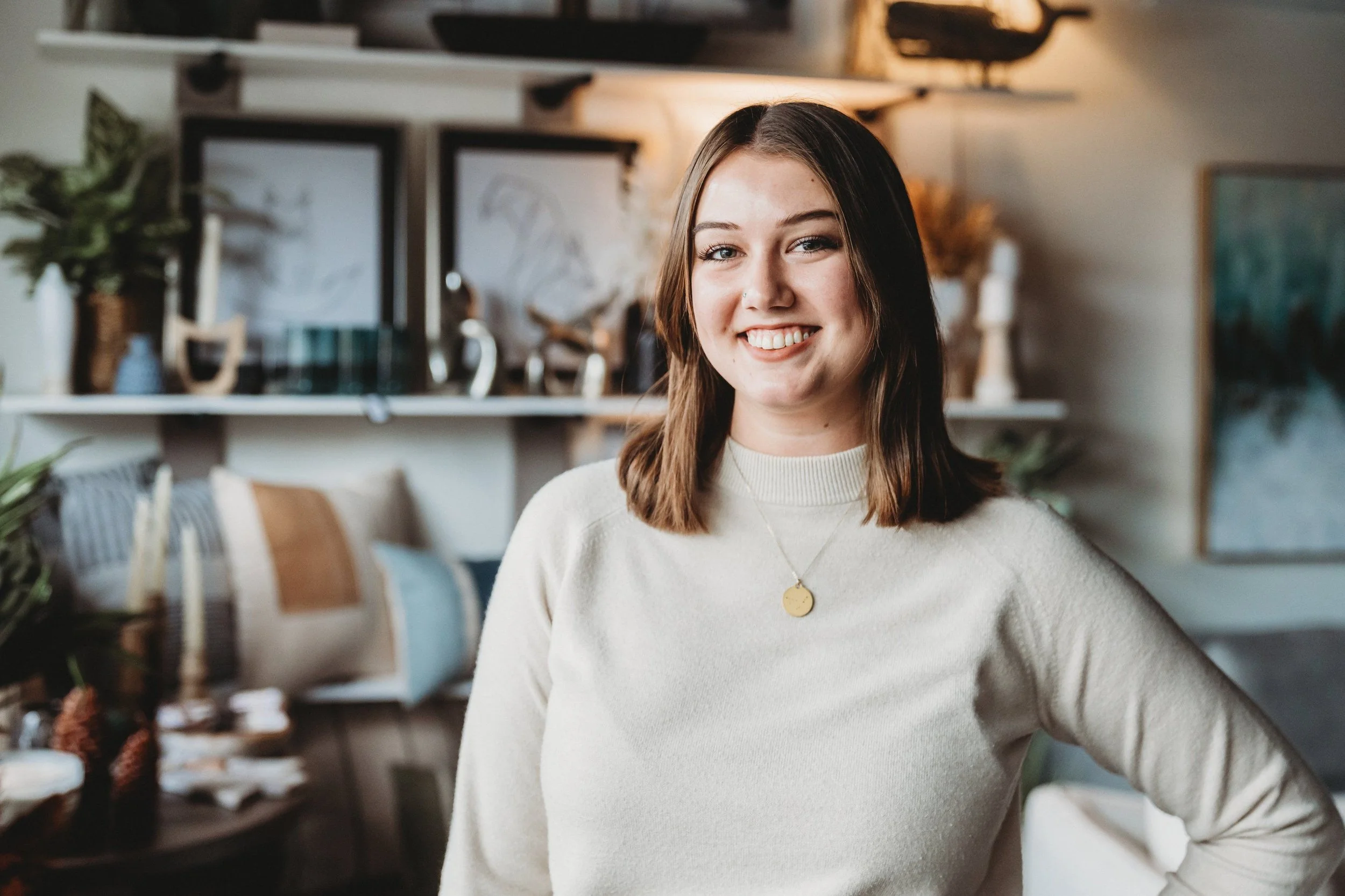 A young woman with shoulder-length brown hair smiling in a cozy, decorated room.