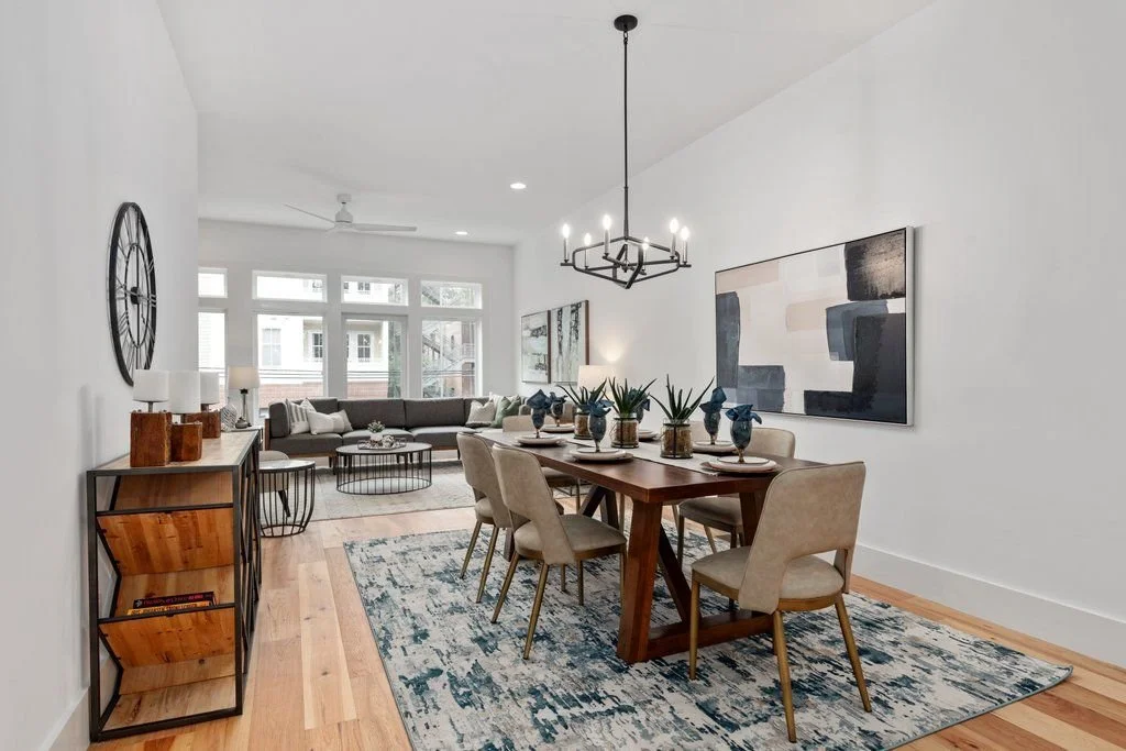 Modern living room and dining area with white walls, hardwood floors, large windows, and minimalist decor. Dining table surrounded by beige chairs with table settings and potted plants, chandelier overhead. Living area with a sectional sofa, coffee tables, and wall art.
