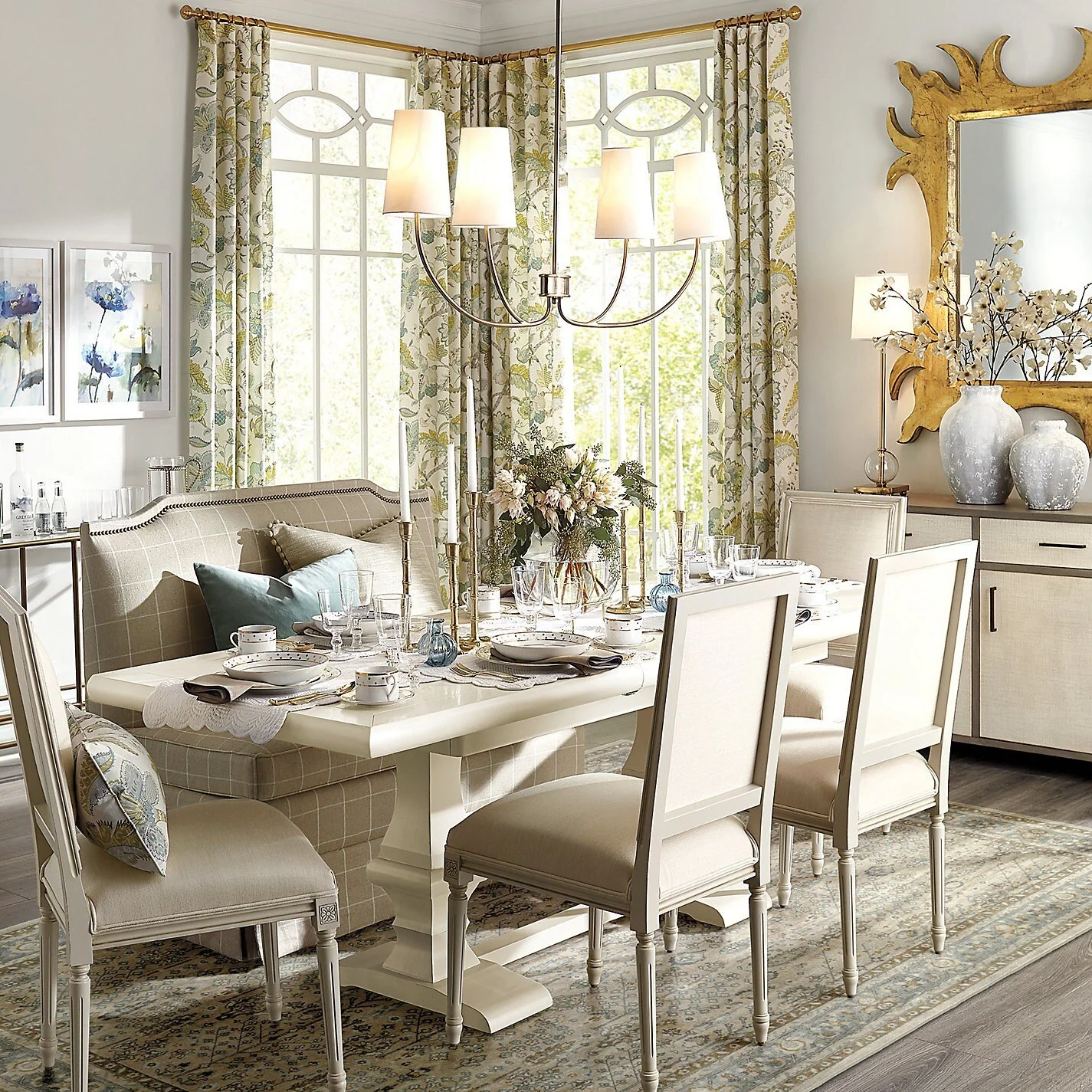 Transitional dining room with a white pedestal table, upholstered dining chairs, built-in banquette seating, floral drapery, sculptural chandelier, gold mirror and layered blue-gray accents.