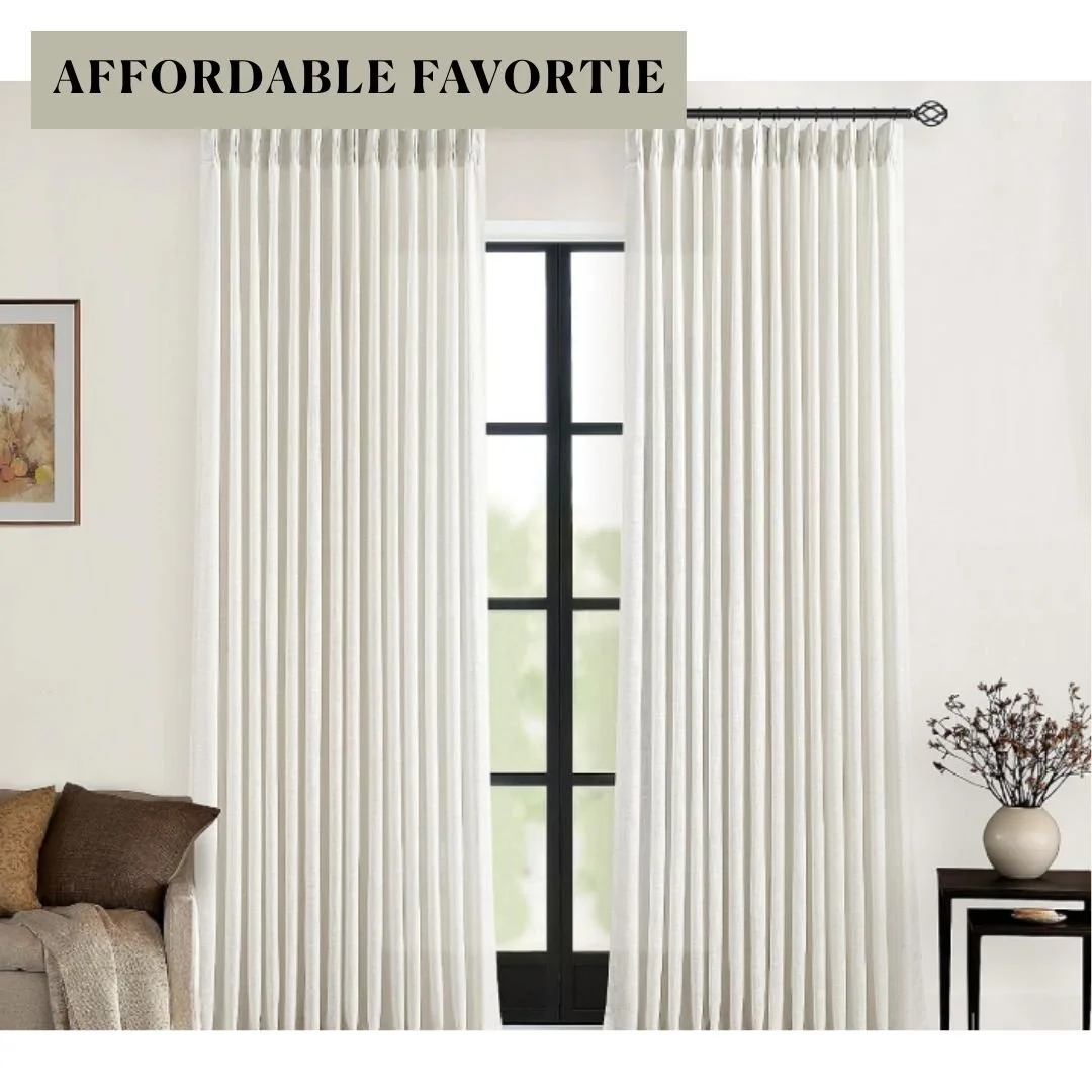 Floor-length ivory linen-look curtains with tailored pinch pleats framing a tall black window in a neutral living room.