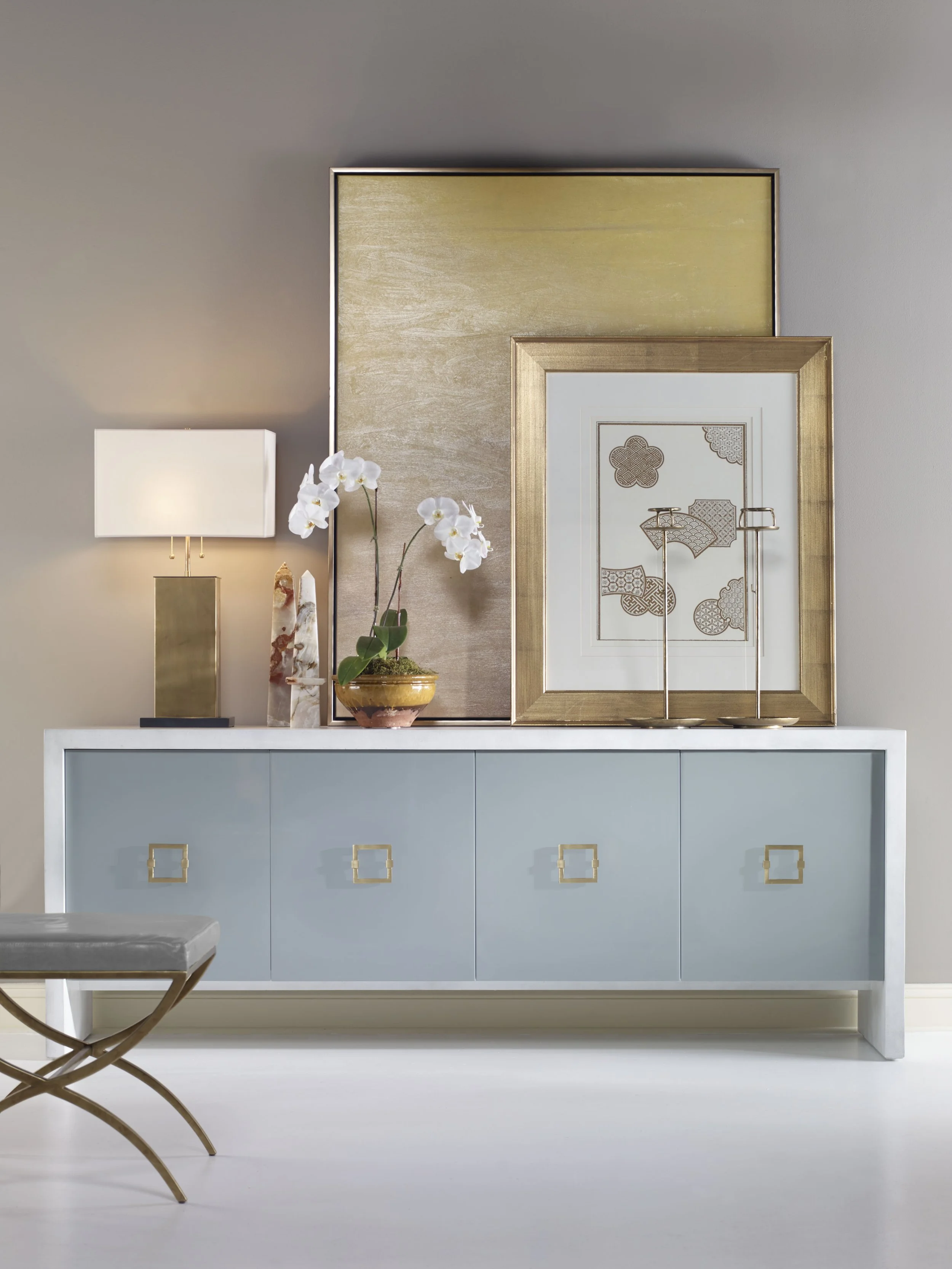 Contemporary foyer vignette featuring a sleek blue-gray credenza with brass hardware, oversized gold-framed artwork, modern table lamp, orchid arrangement, and metallic accents, creating a sophisticated and polished contemporary interior.