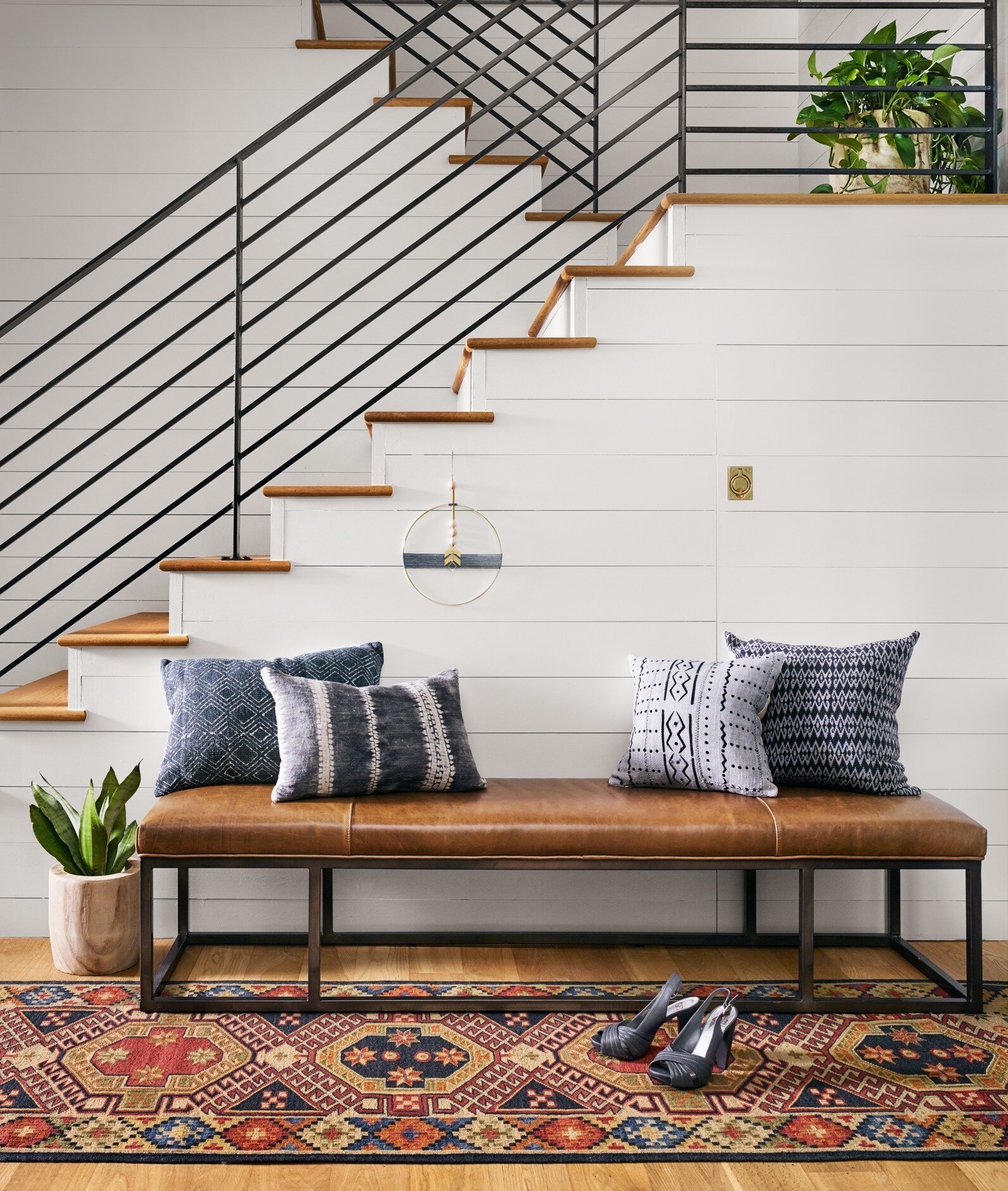 Modern rustic foyer with white plank walls, wood stairs, black metal railing and a leather bench with patterned pillows.