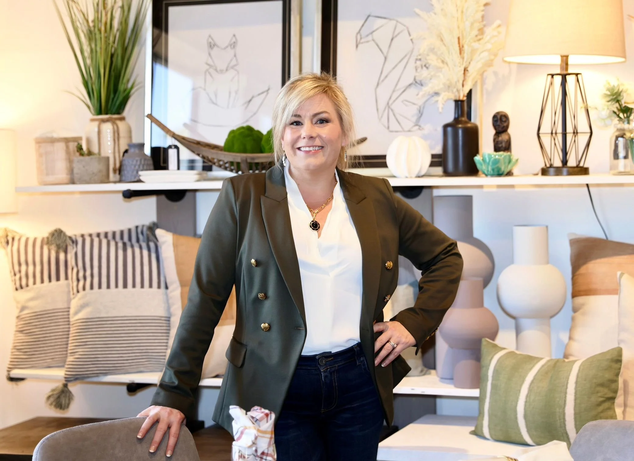 A woman with short blonde hair smiling in a living room, standing with her hand on the back of a chair. The room has decorative pillows, artwork, and vases on shelves.