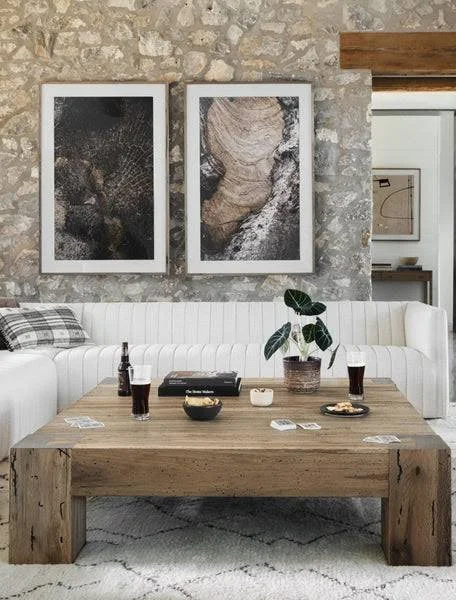 Modern rustic dining area featuring a reclaimed wood dining table, curved white upholstered banquette seating, stone accent walls, oversized abstract artwork, exposed wood beams, and layered natural textures in a neutral color palette.