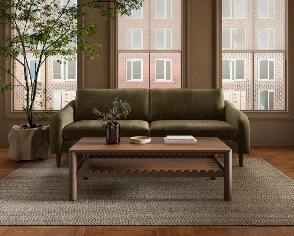 Scandinavian and Mid-Century Modern living room featuring an olive green sofa with tapered wood legs, a warm wood coffee table, textured neutral area rug, natural greenery, and large windows that create a clean, functional, and inviting space.