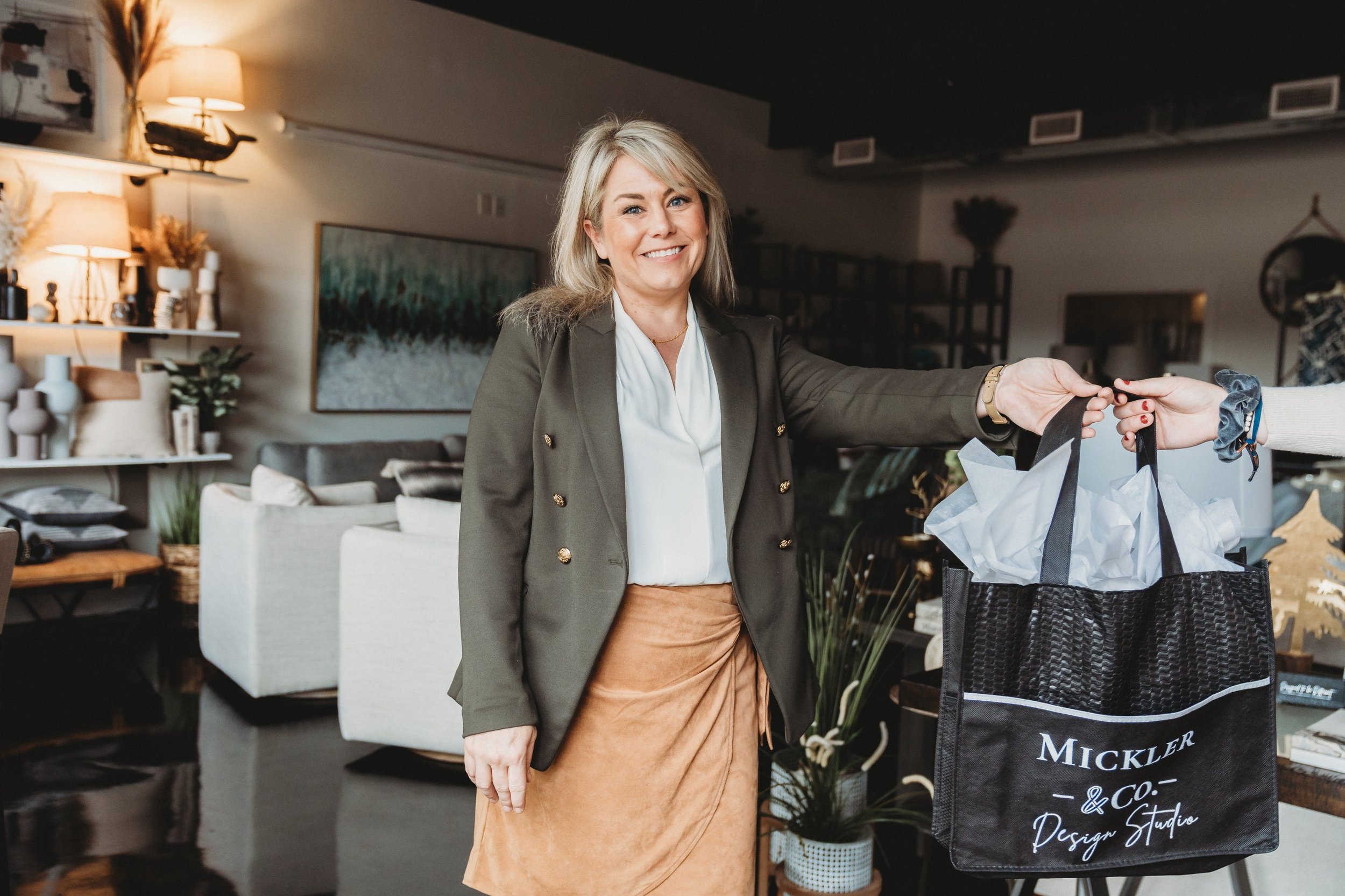 A smiling woman with shoulder-length blonde hair handing over a gift bag to someone in a cozy, well-decorated living room.