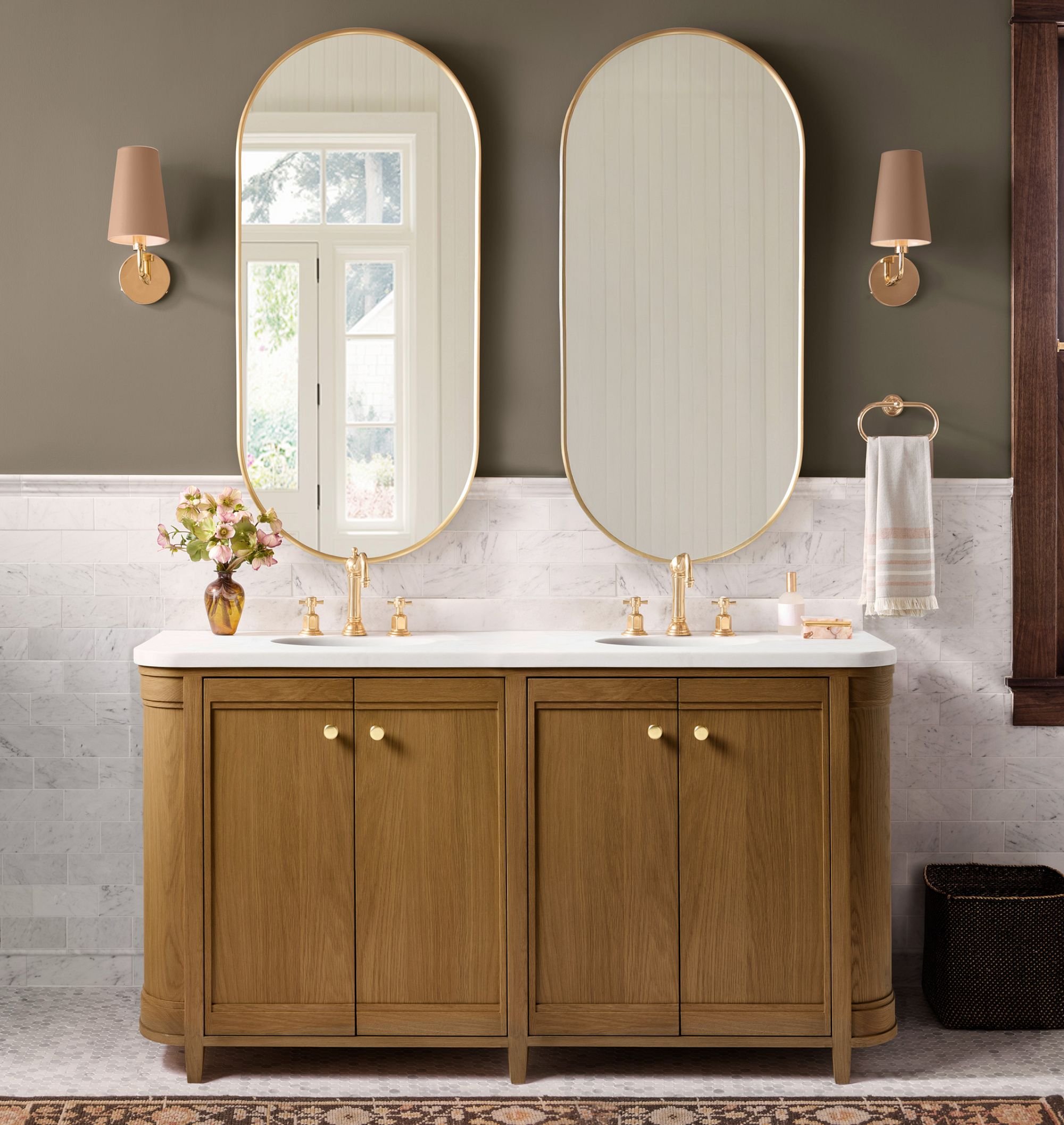Transitional bathroom with a white double vanity, warm wood cabinetry, brass fixtures, arched mirrors, and marble tile walls.