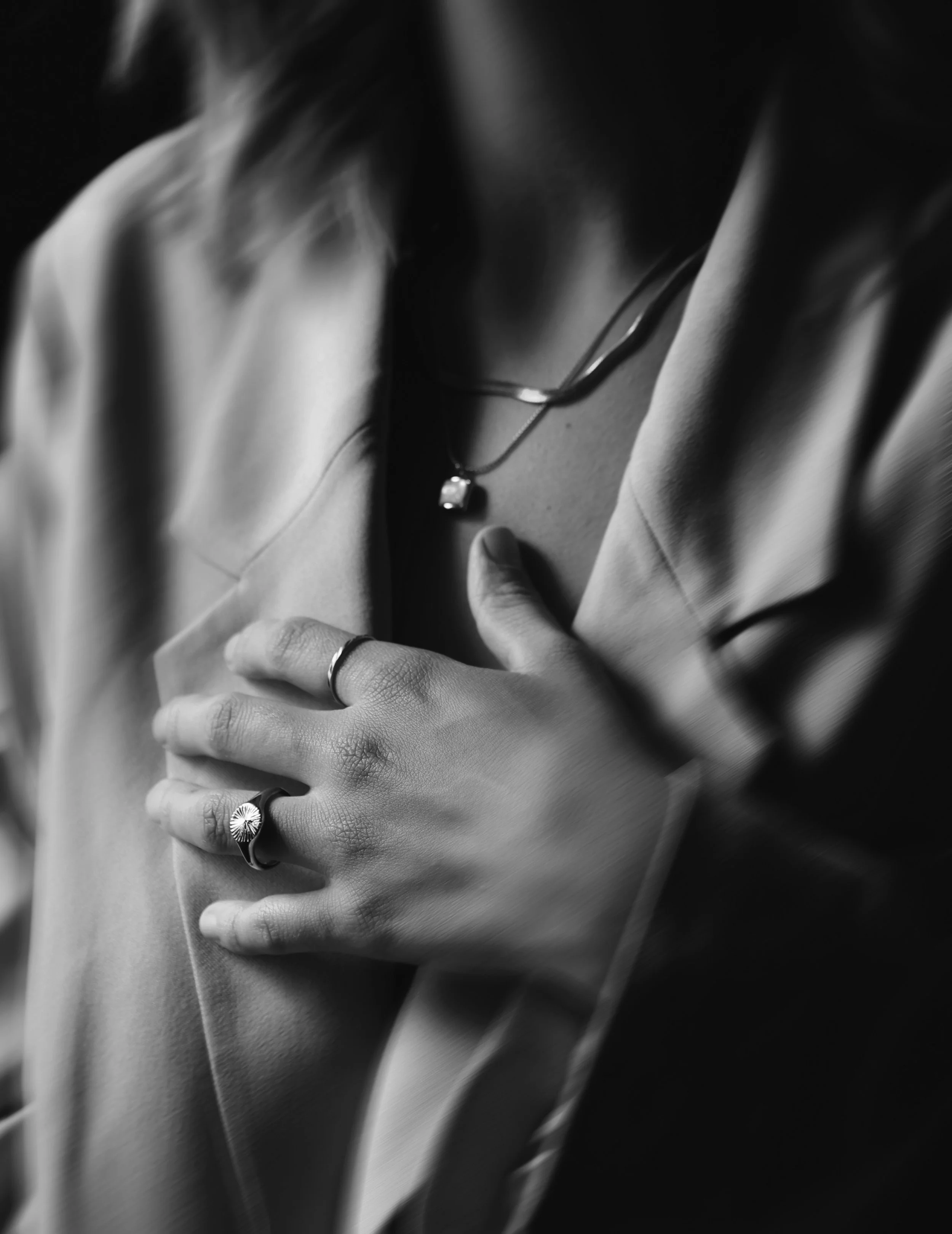 Close-up black and white photo of a person's hand with rings touching their chest, wearing a suit with a necklace featuring a cubic zirconia pendant.