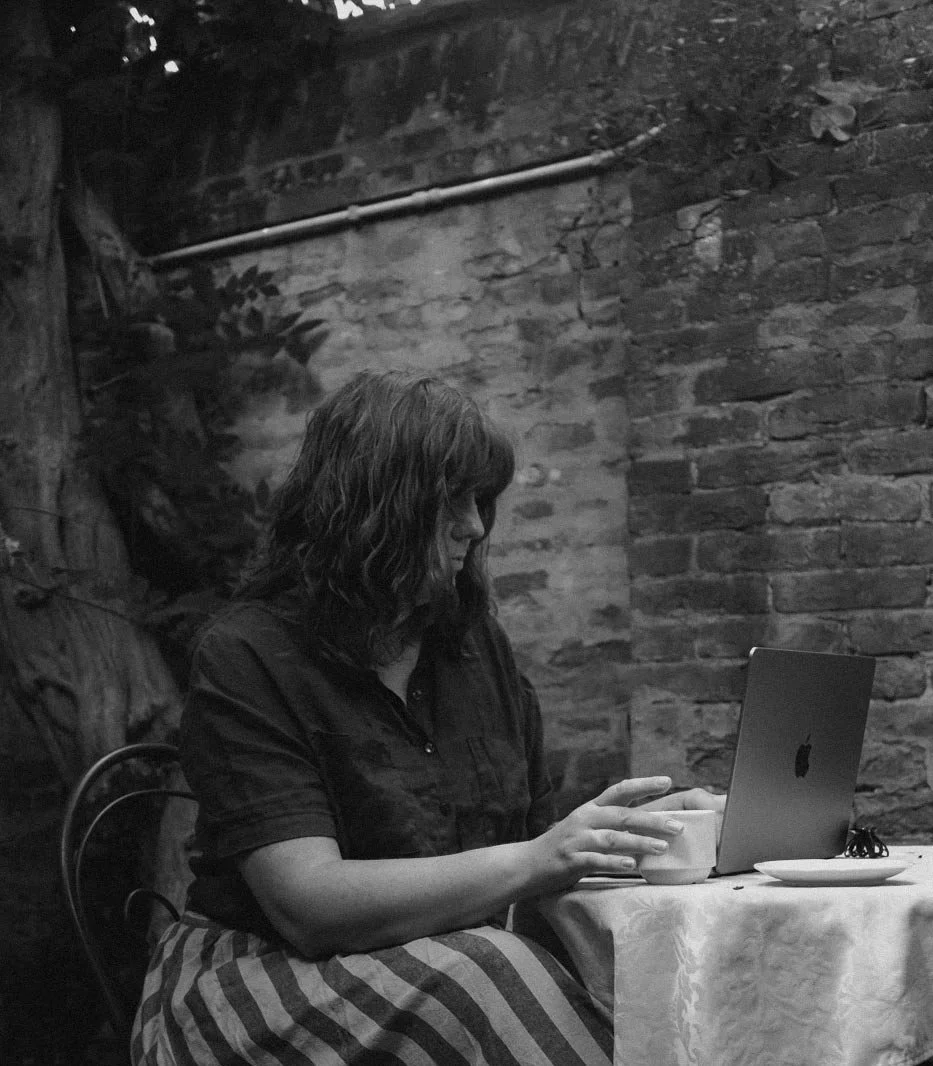 A woman with shoulder-length curly hair sitting at a table with a laptop, cup, and plate in front of her against a brick wall background.
