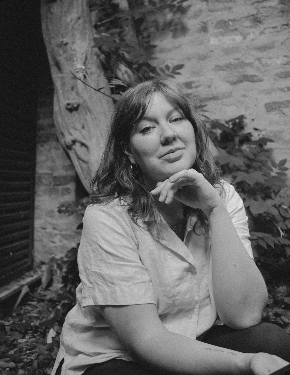 A black and white photo of a woman with shoulder-length wavy hair, wearing a short-sleeved shirt, sitting outdoors near a brick wall and leafy plants, resting her chin on her left hand and looking at the camera.