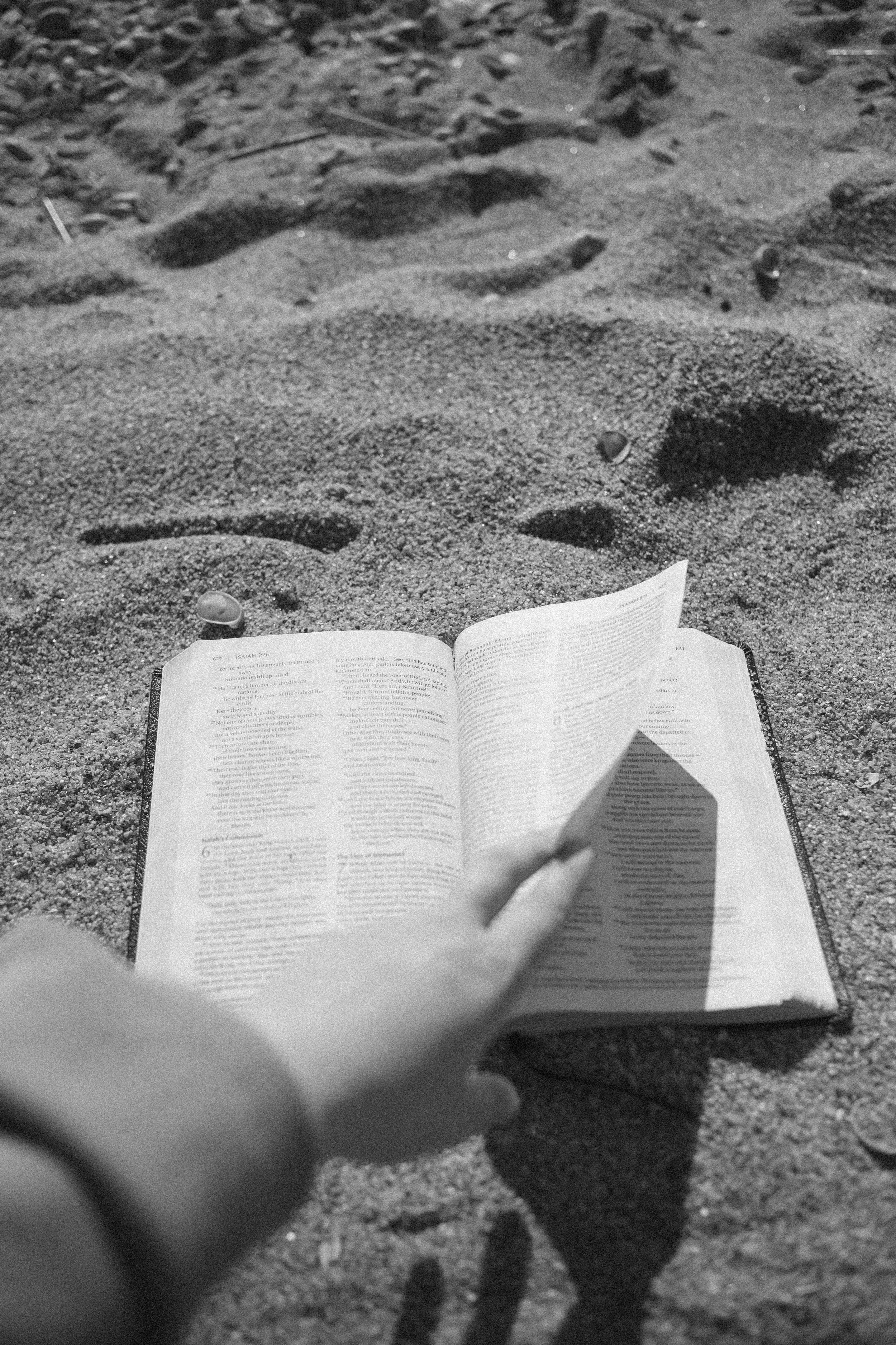 Black and white photo of a person holding an open book on a sandy beach, with a visible shadow of the person cast on the sand.