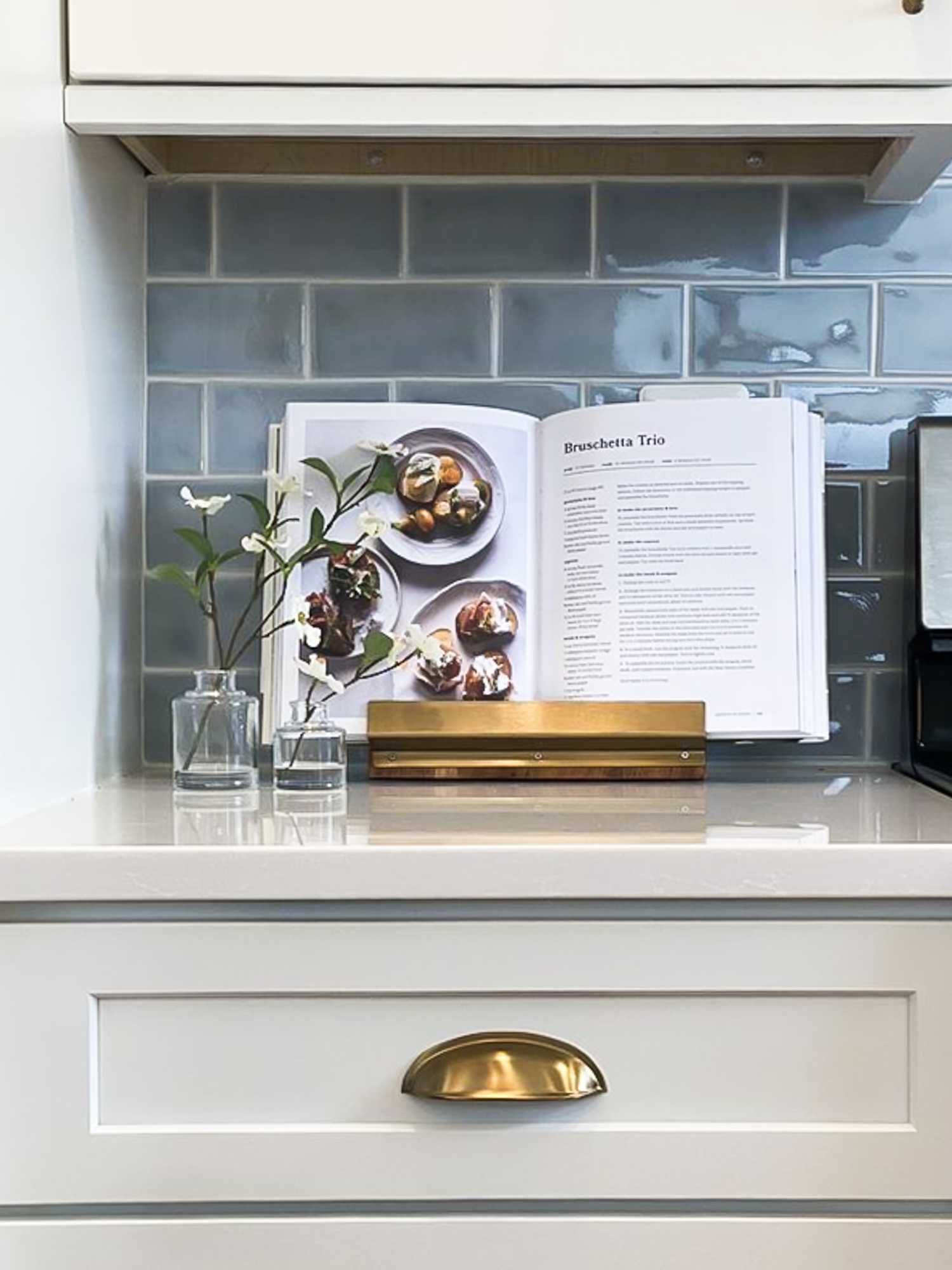 Open cookbook on a kitchen counter, displaying a recipe for Bruschetta Trio, with a glass vase of flowers and two glasses of water nearby.