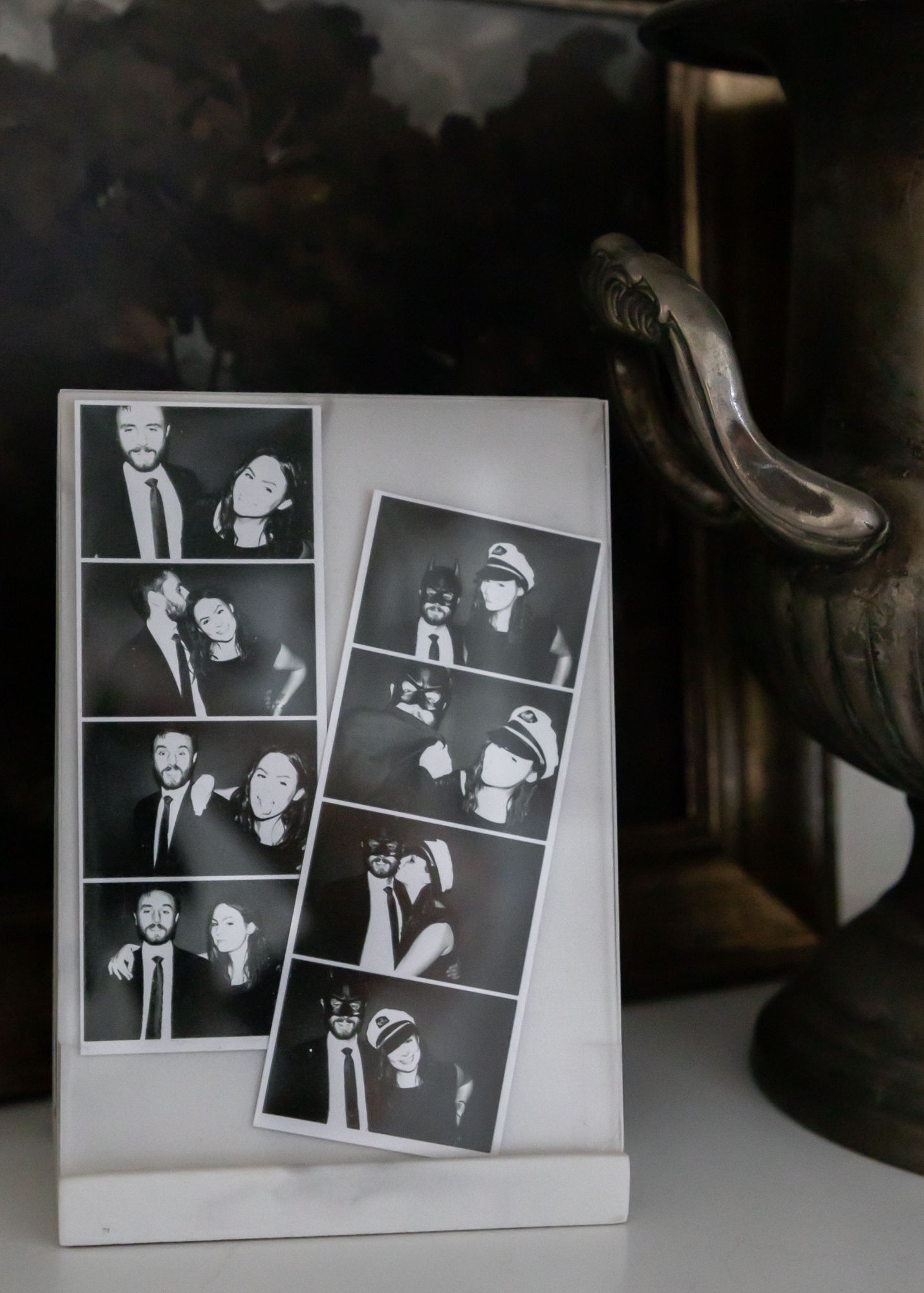Black and white photo booth strip with four pictures of a man and woman, and a second photo strip with four images of the same woman and a man wearing various costumes, such as a superhero mask and hat. The strips are held in a white holder and are placed on a table next to a dark decorative object or furniture.