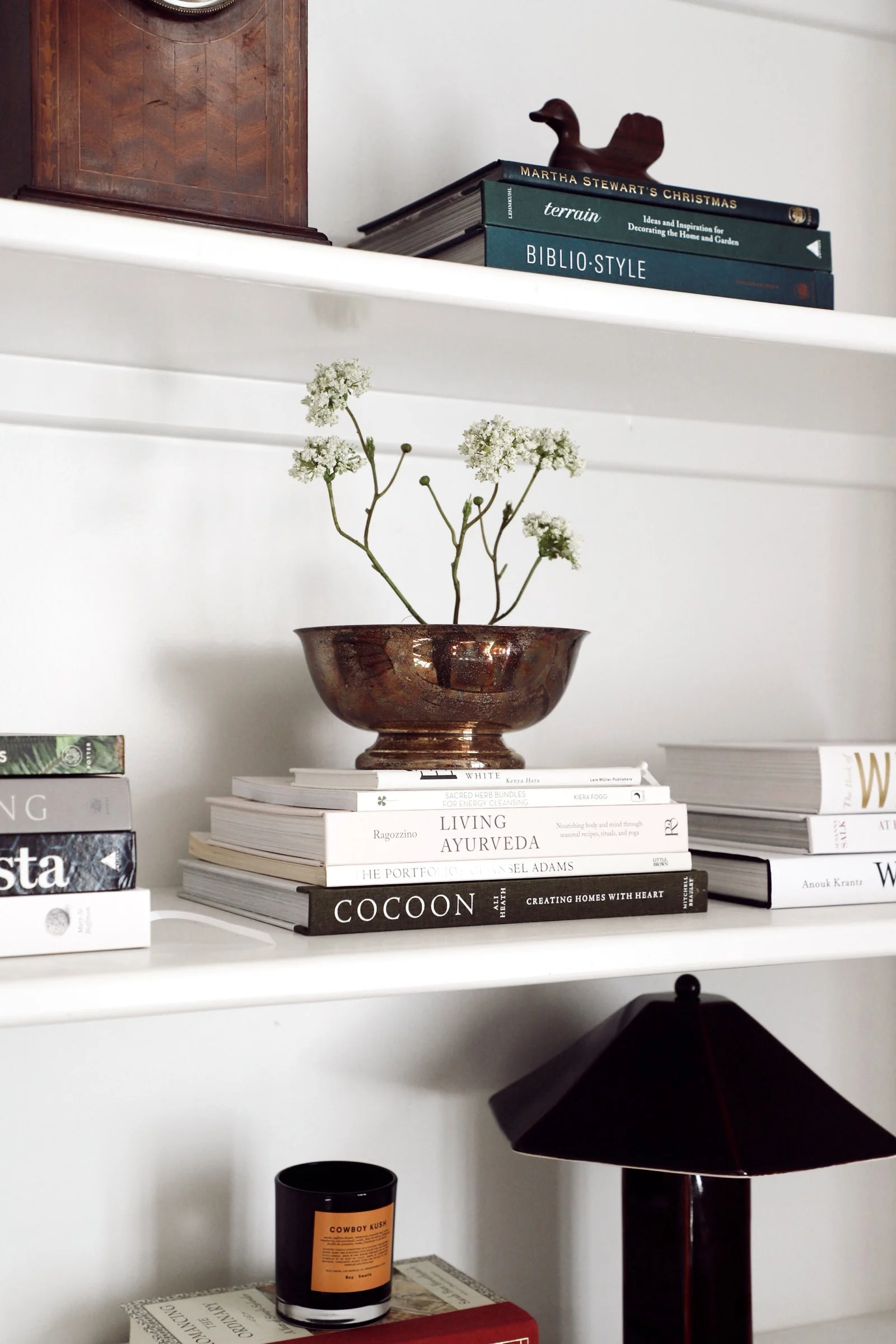 A white shelf holding books, a decorative brown and metallic bowl with white flowers, a small black lamp with a conical shade, and a candle.