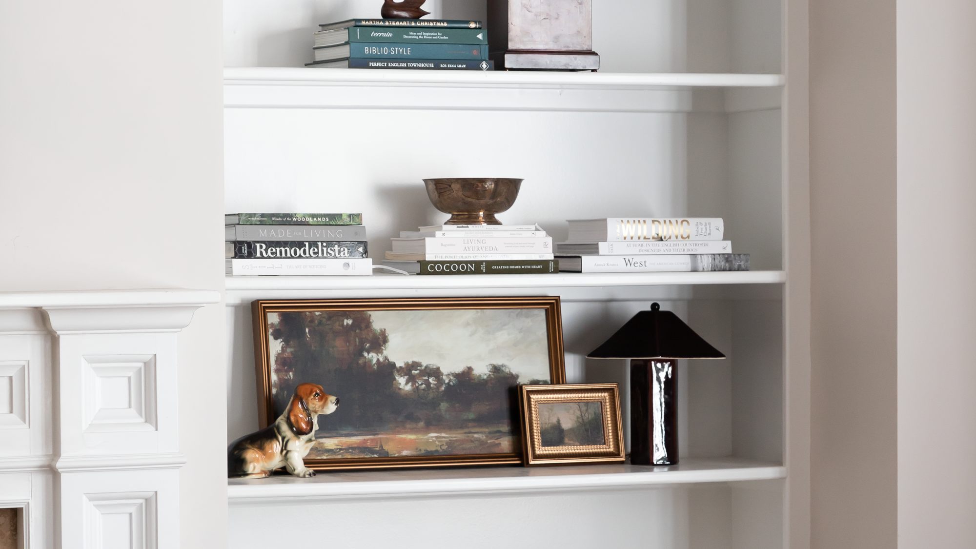 White built-in bookshelf with books, framed landscape painting, a small picture frame, a black table lamp, and a decorative ceramic dog figurine.