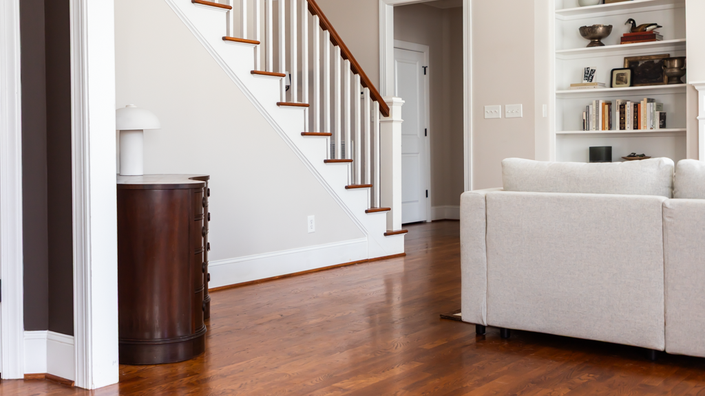 Living room with wooden floors, a beige sofa, a wooden staircase with white risers, and built-in bookshelves filled with books and decorative items.