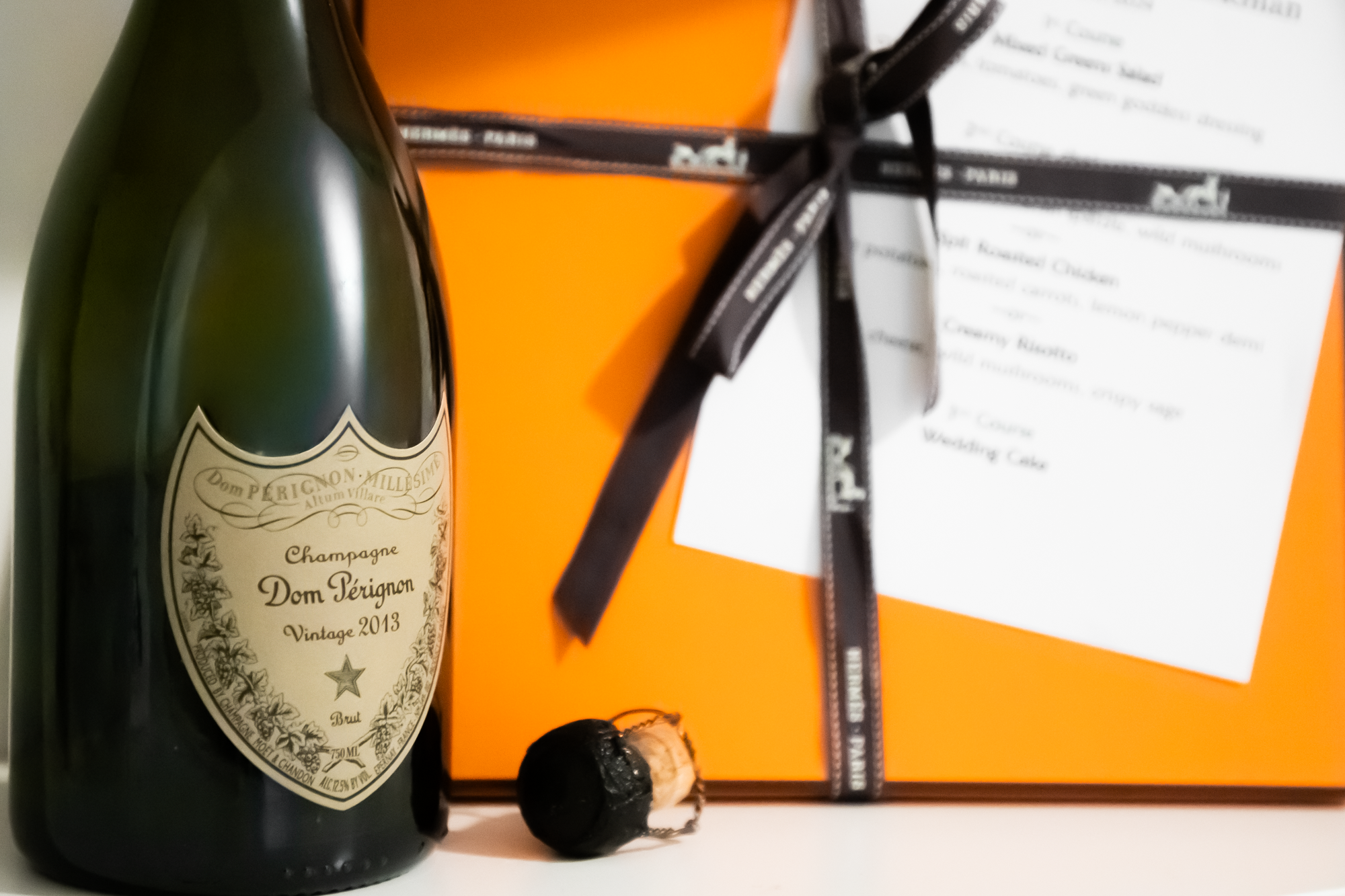 A bottle of Dom Pérignon Vintage 2013 champagne, a champagne cork with a wire cage, and an orange gift box with a black ribbon and a white card.