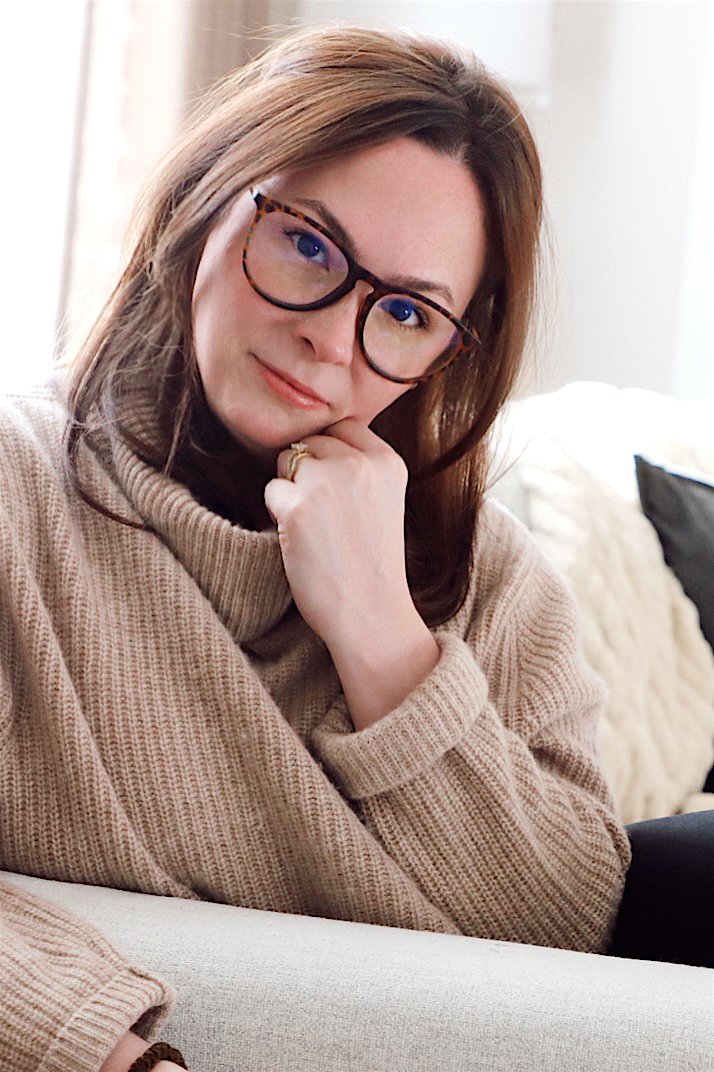 Lauren Murphy- a woman with brown hair, wearing glasses and a beige turtleneck sweater, sitting on a sofa with a thoughtful expression, resting her chin on her hand.