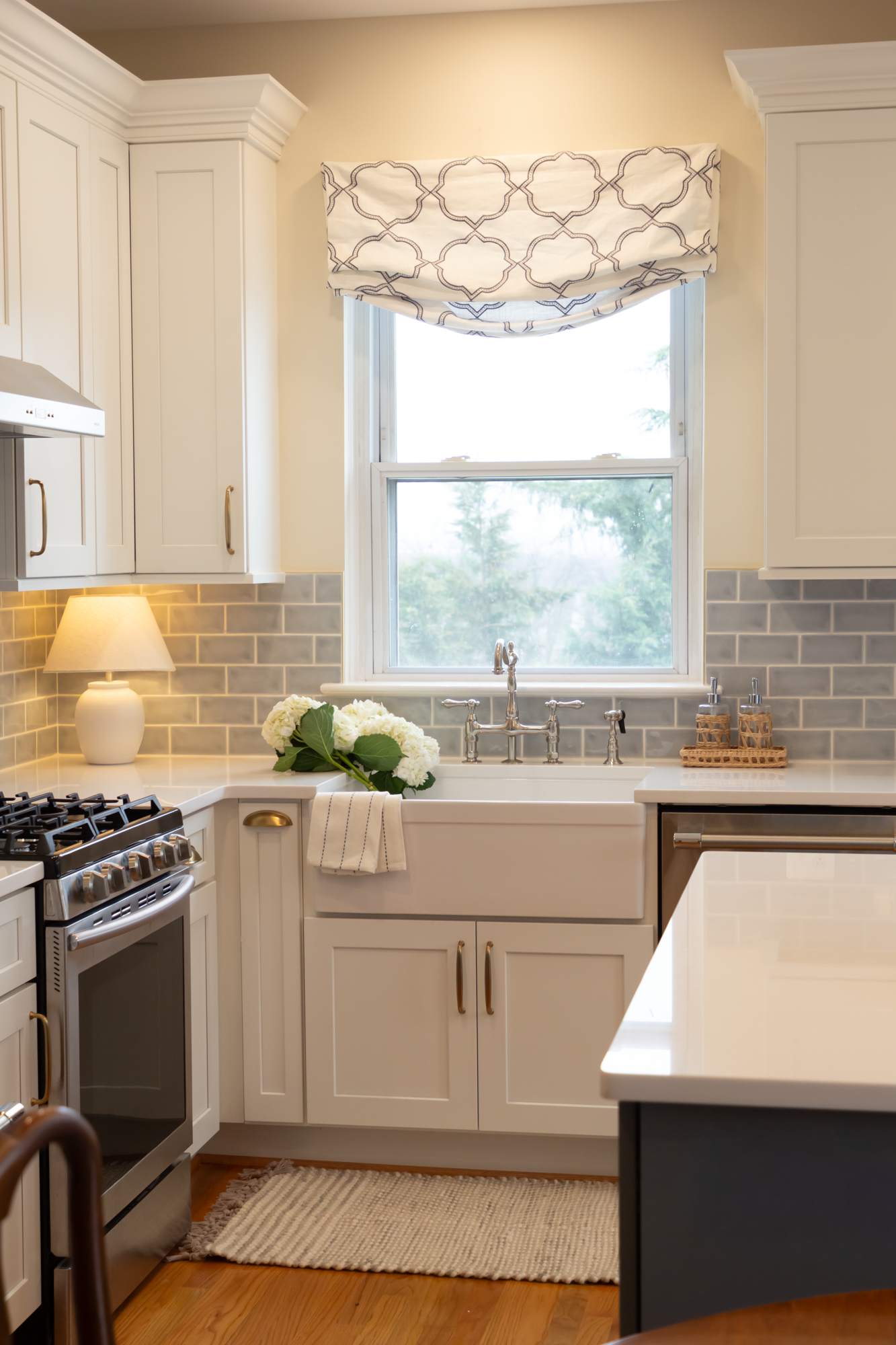 Bright kitchen with white cabinets, a window with a patterned valance, a farmhouse sink with a bouquet of white flowers, a beige rug, and a gas stove.