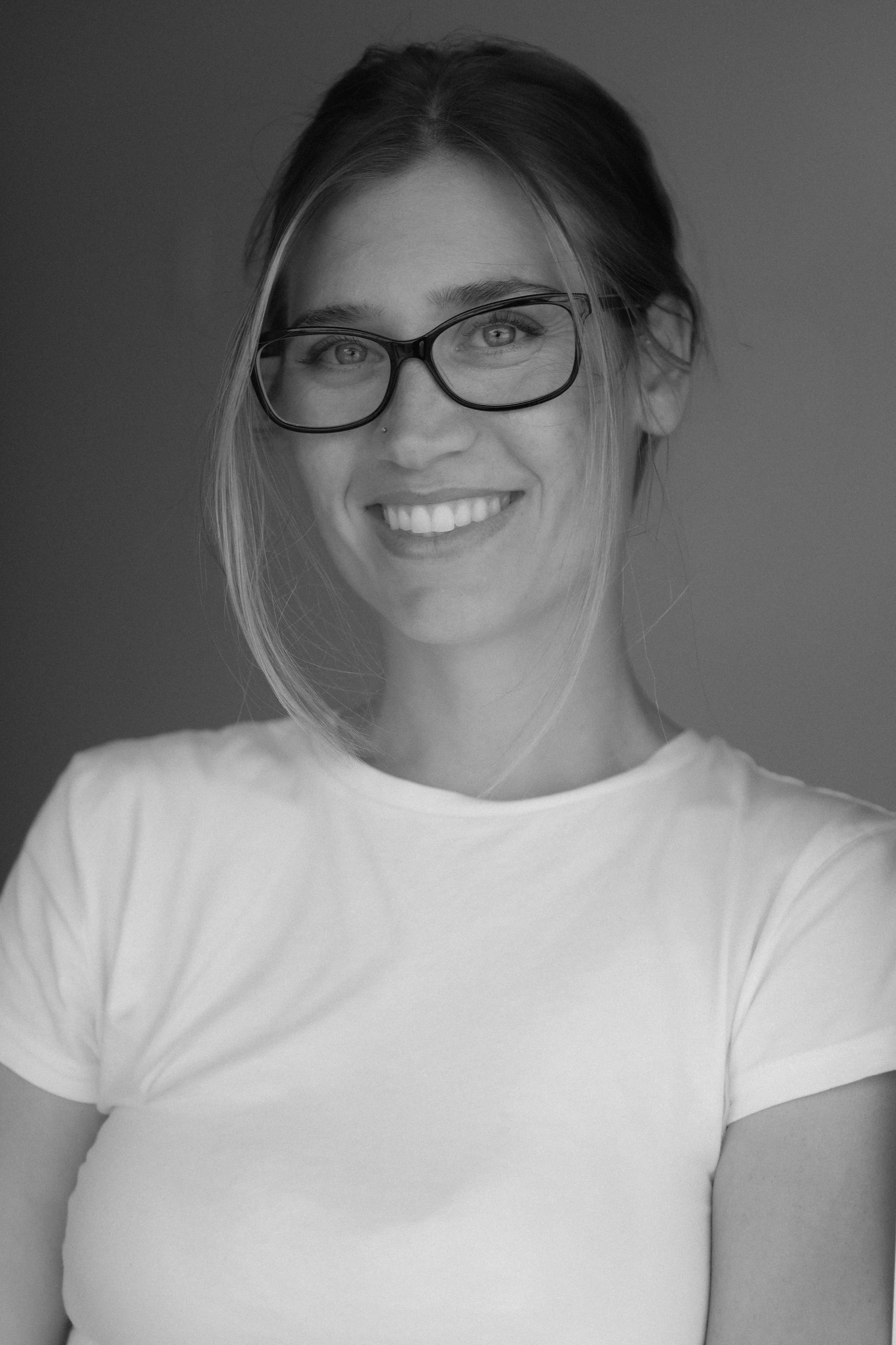 A smiling woman with glasses and a nose piercing wearing a light-colored t-shirt, against a plain background.