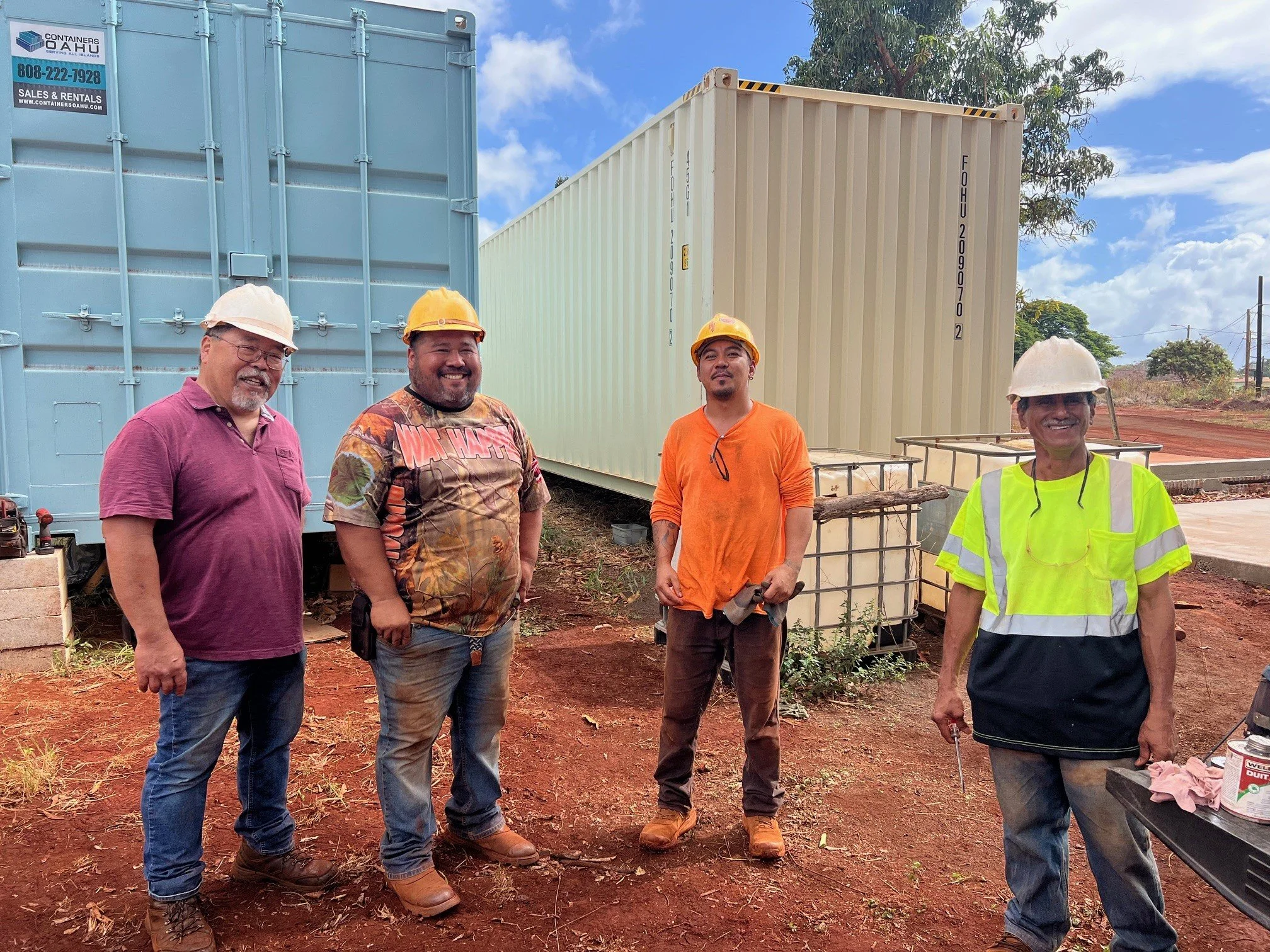 Four men wearing construction helmets and safety vests standing outdoors near shipping containers, with a partly cloudy sky and trees in the background.