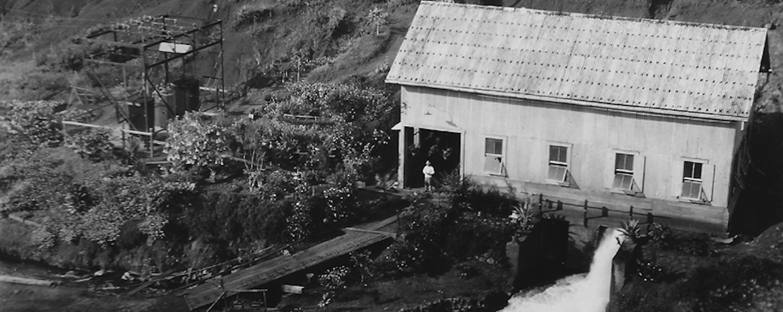 Black and white aerial view of a rural scene with a wooden house, garden, and a watermill near a small waterfall.
