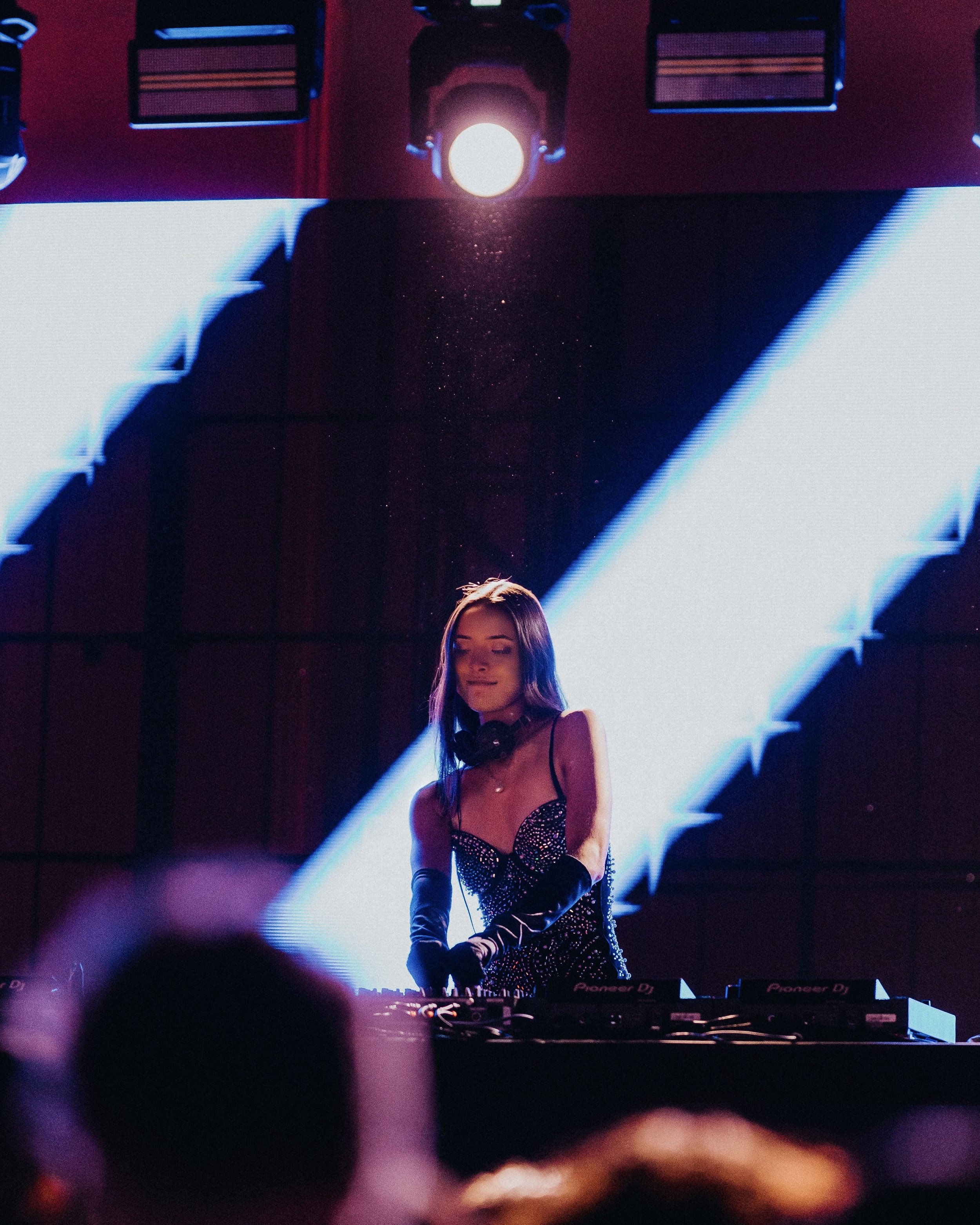 A female DJ with long hair, wearing a black and silver dress and black gloves, performing at a party or concert with a bright light and large screen in the background.
