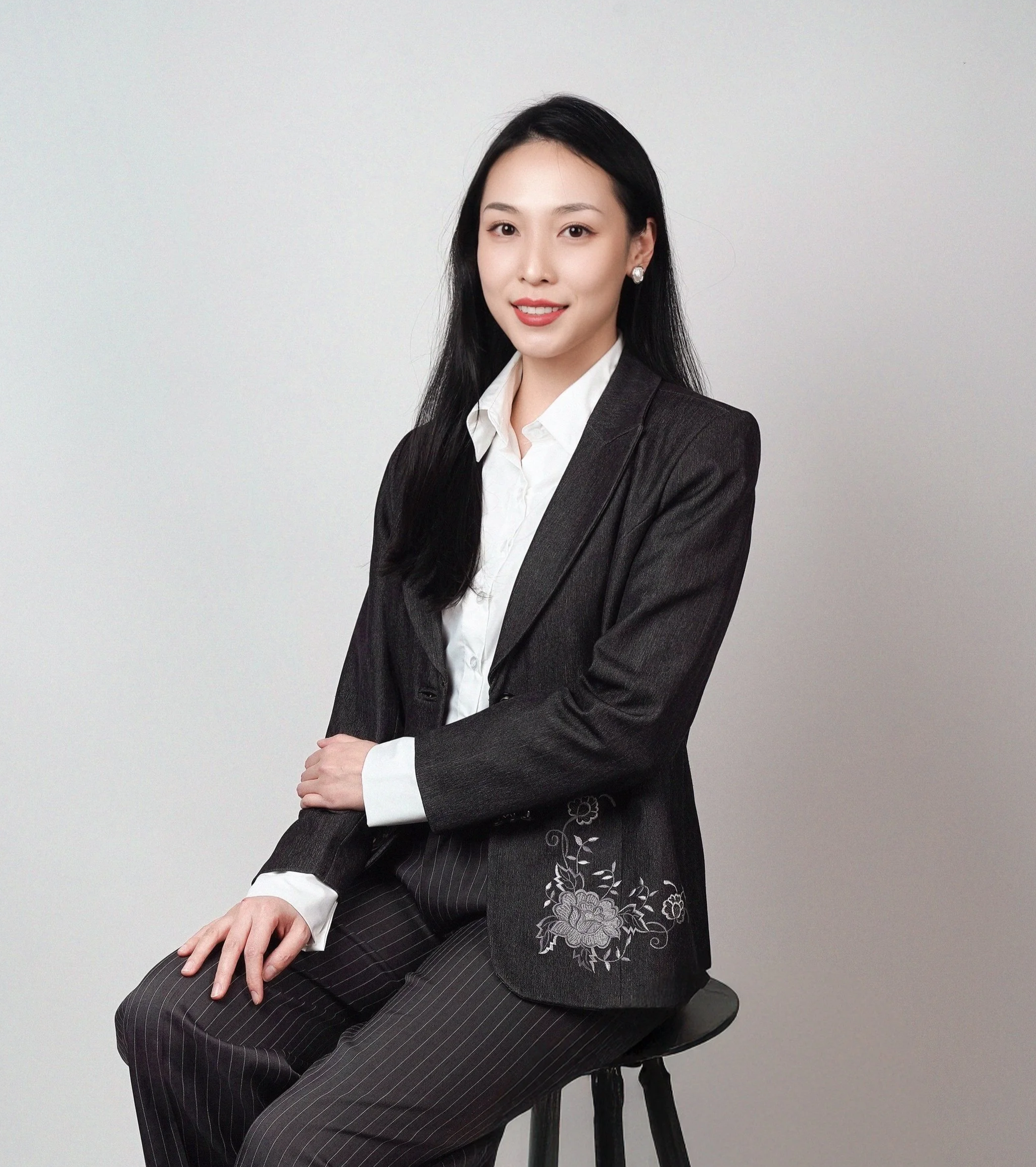 A young woman with long black hair, wearing a dark gray pinstripe suit with floral embroidery, sitting on a black stool against a plain light background.