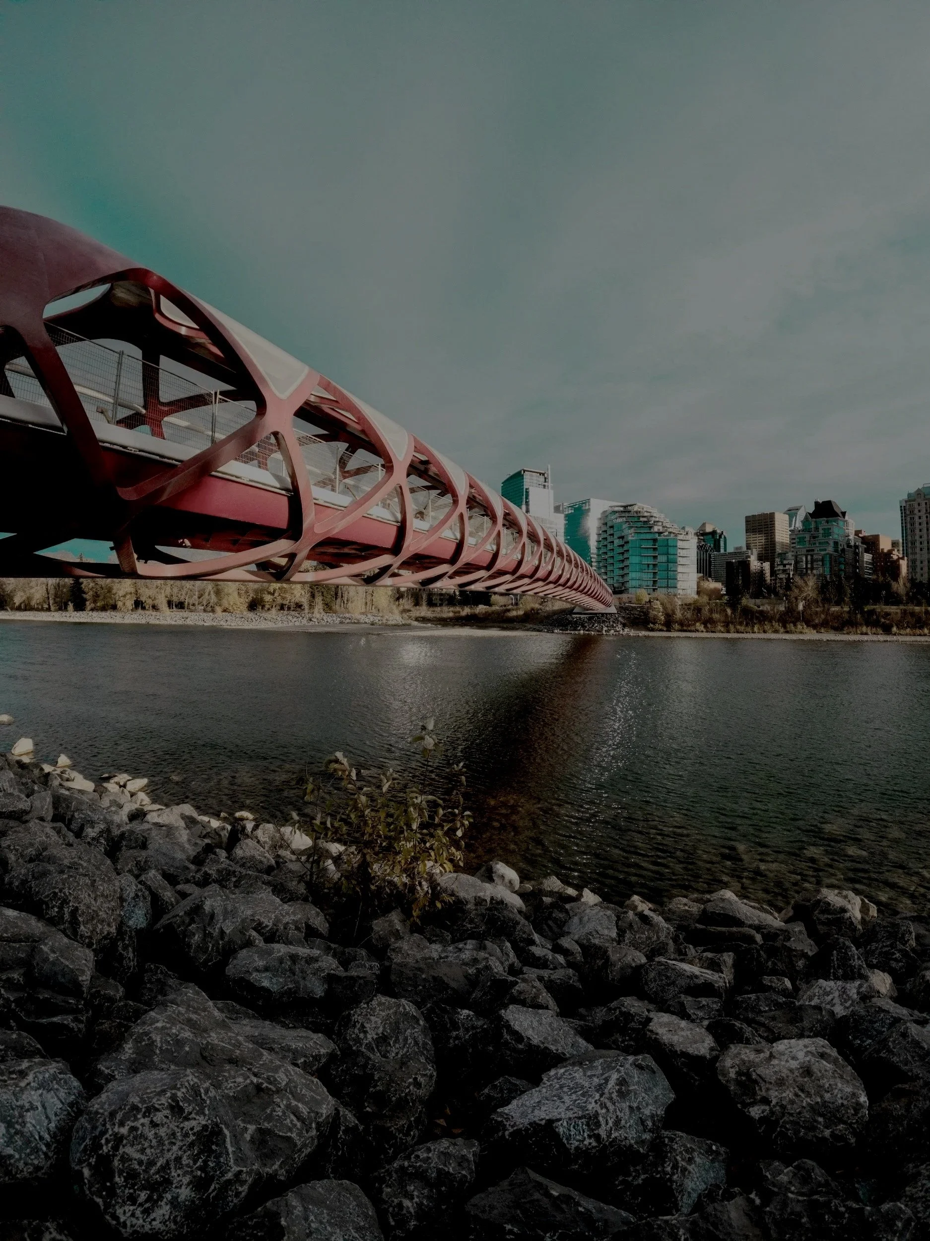 A modern cityscape with a distinctive red bridge extending over a body of water, with a skyline of high-rise buildings and a cloudy sky in the background.