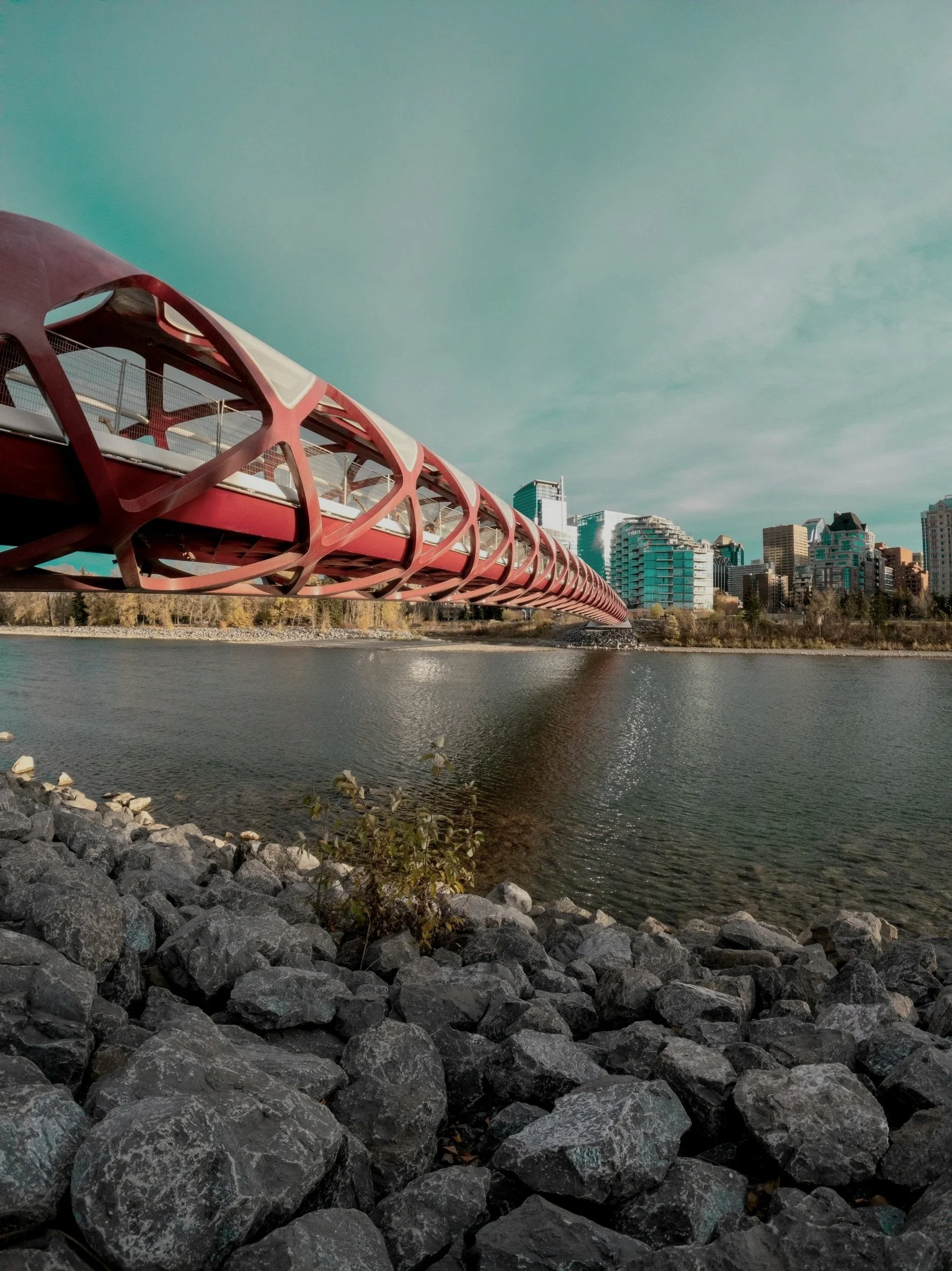 A modern, red pedestrian bridge over a body of water with city buildings in the background and rocks along the shore.