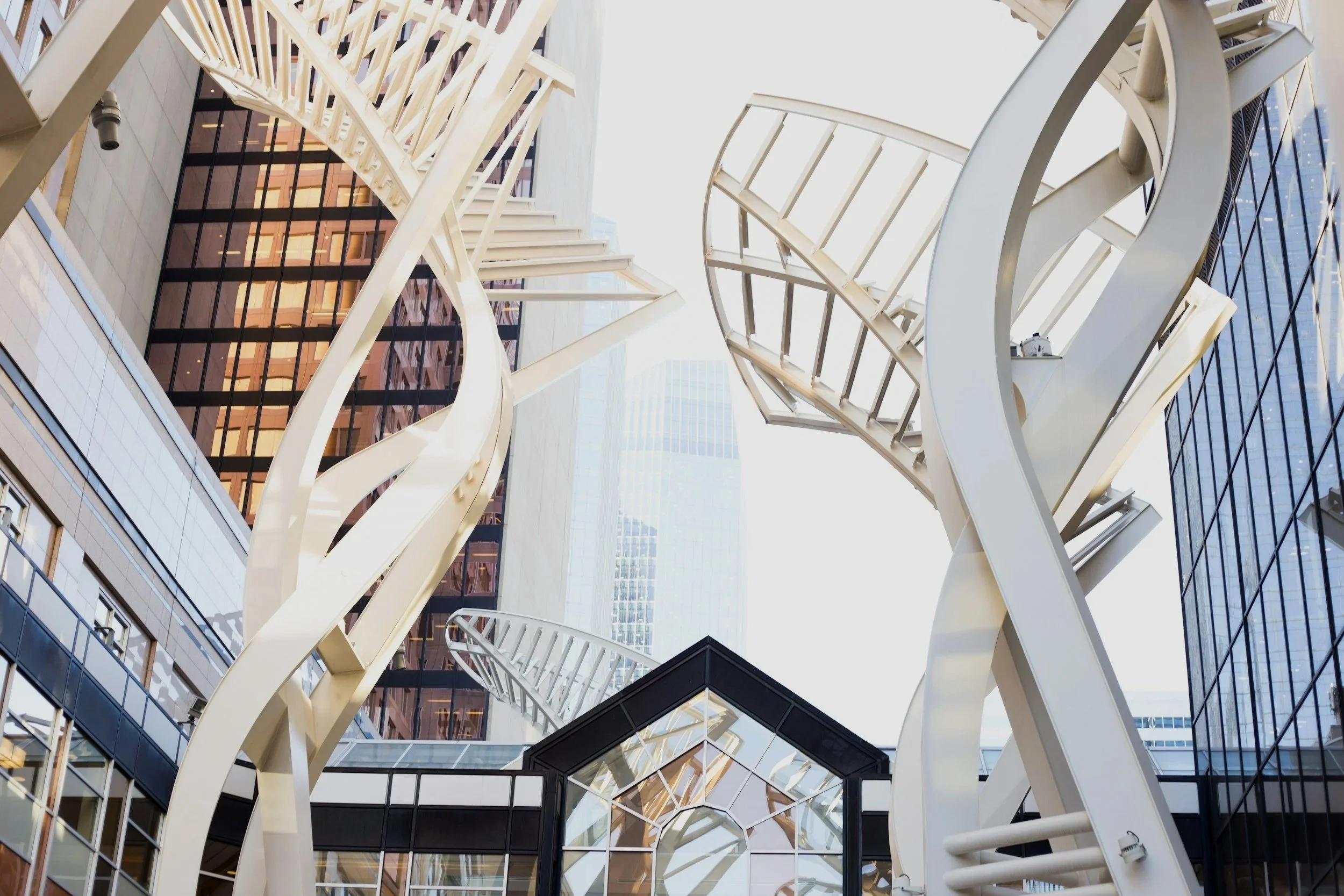 Low-angle view of modern architectural sculptures with twisting, spiral, and ladder-like elements in an urban setting, surrounded by glass buildings.