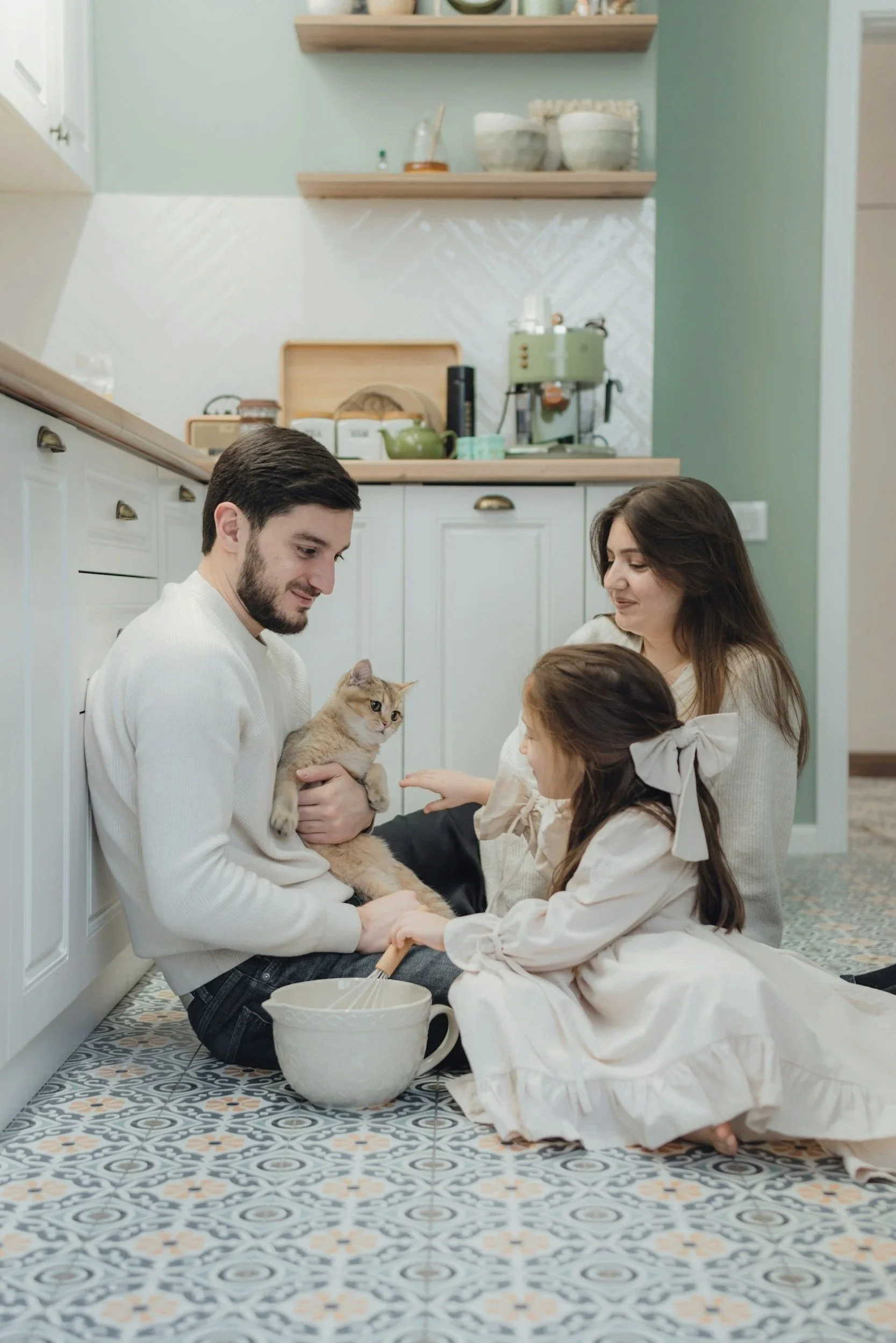 Family of three in kitchen with cat, girl reaching towards cat, man holding cat, woman watching, patterned floor, green wall, kitchen shelves and appliances in background.