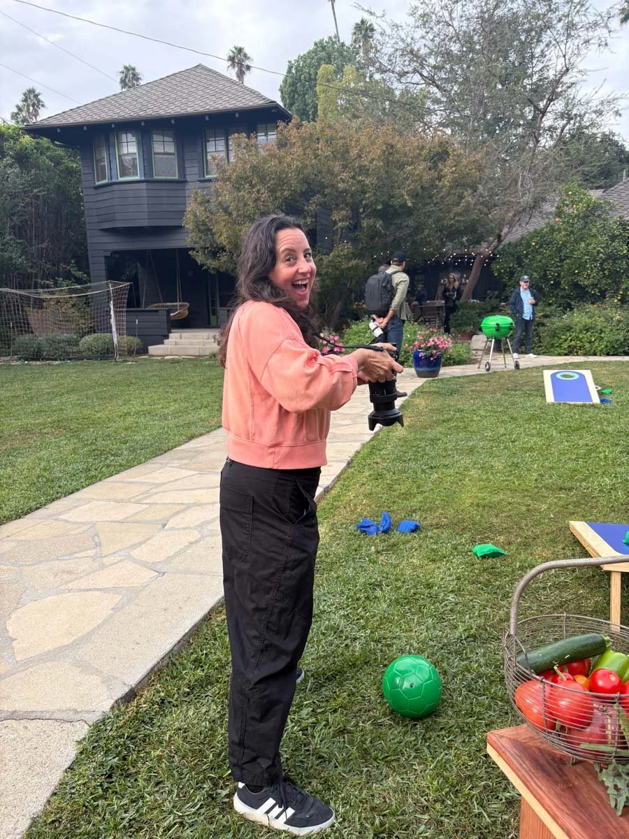 A woman smiling and holding a camera stands on a grassy backyard with a stone pathway, with a house and other people in the background. There is a green ball, a basket of vegetables, and cornhole game boards nearby.
