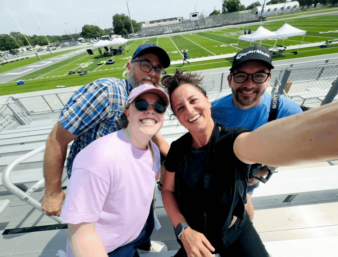 Group of four smiling people taking a selfie on bleachers at a sports stadium during the day, with a football field in the background.