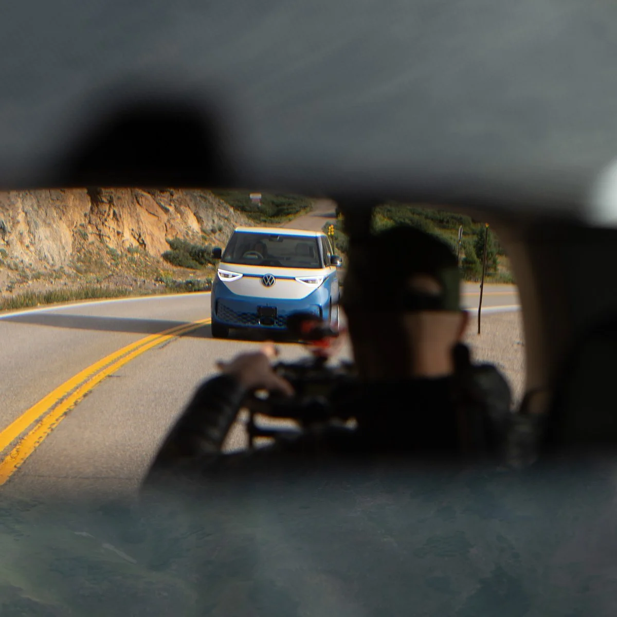 A view from inside a vehicle looking through the rearview mirror at a blue and white Volkswagen ID. Buzz electric van on a winding mountain road.