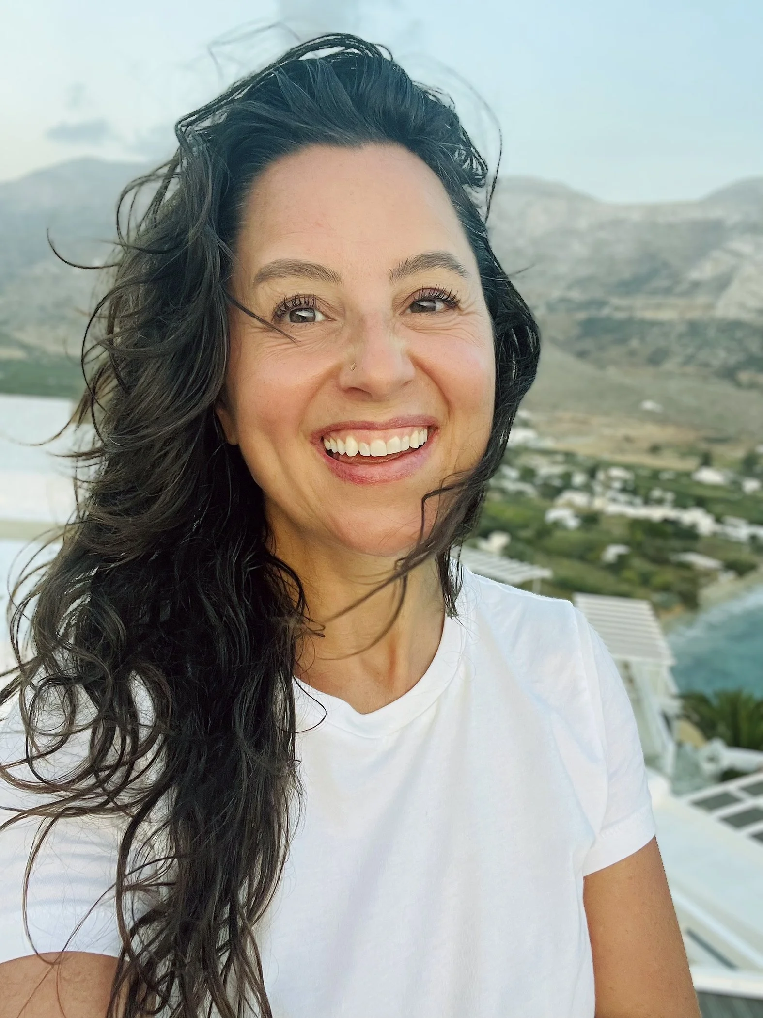 A woman with long, wavy black hair smiling at the camera with a background of mountains, a green landscape, and a coastline.
