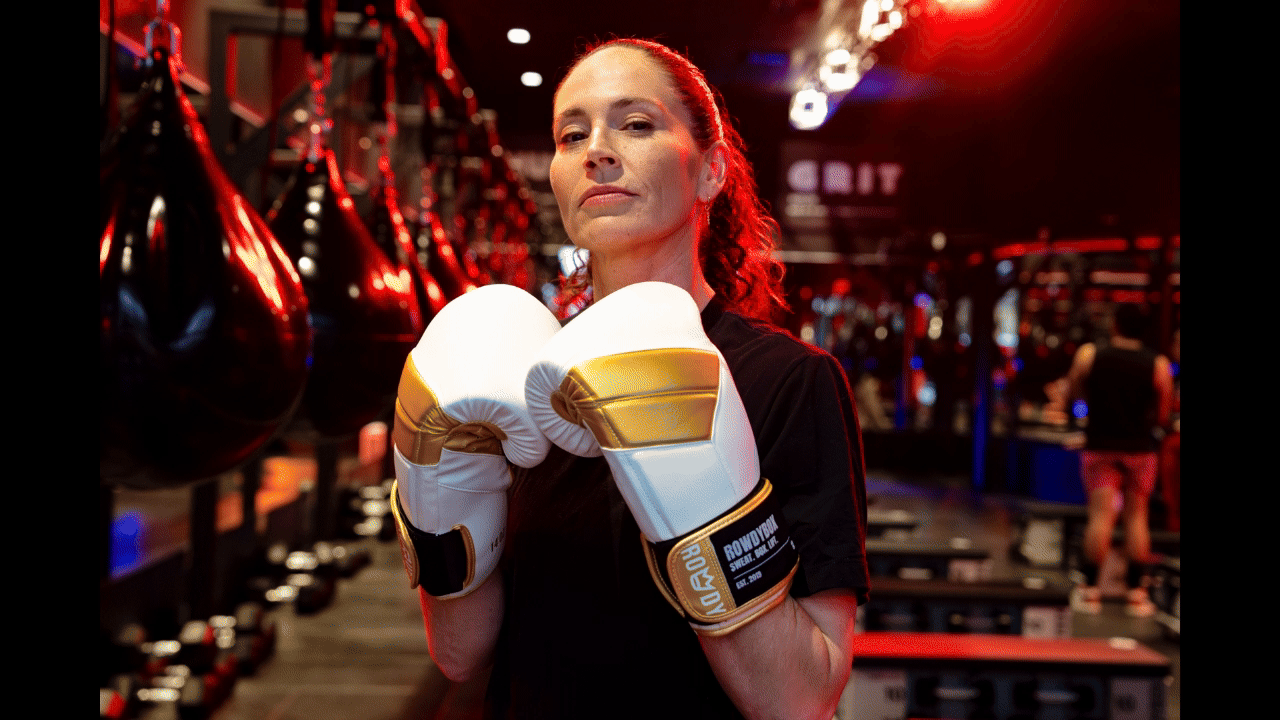 A woman in boxing gloves standing in a boxing gym with punching bags hanging in the background.
