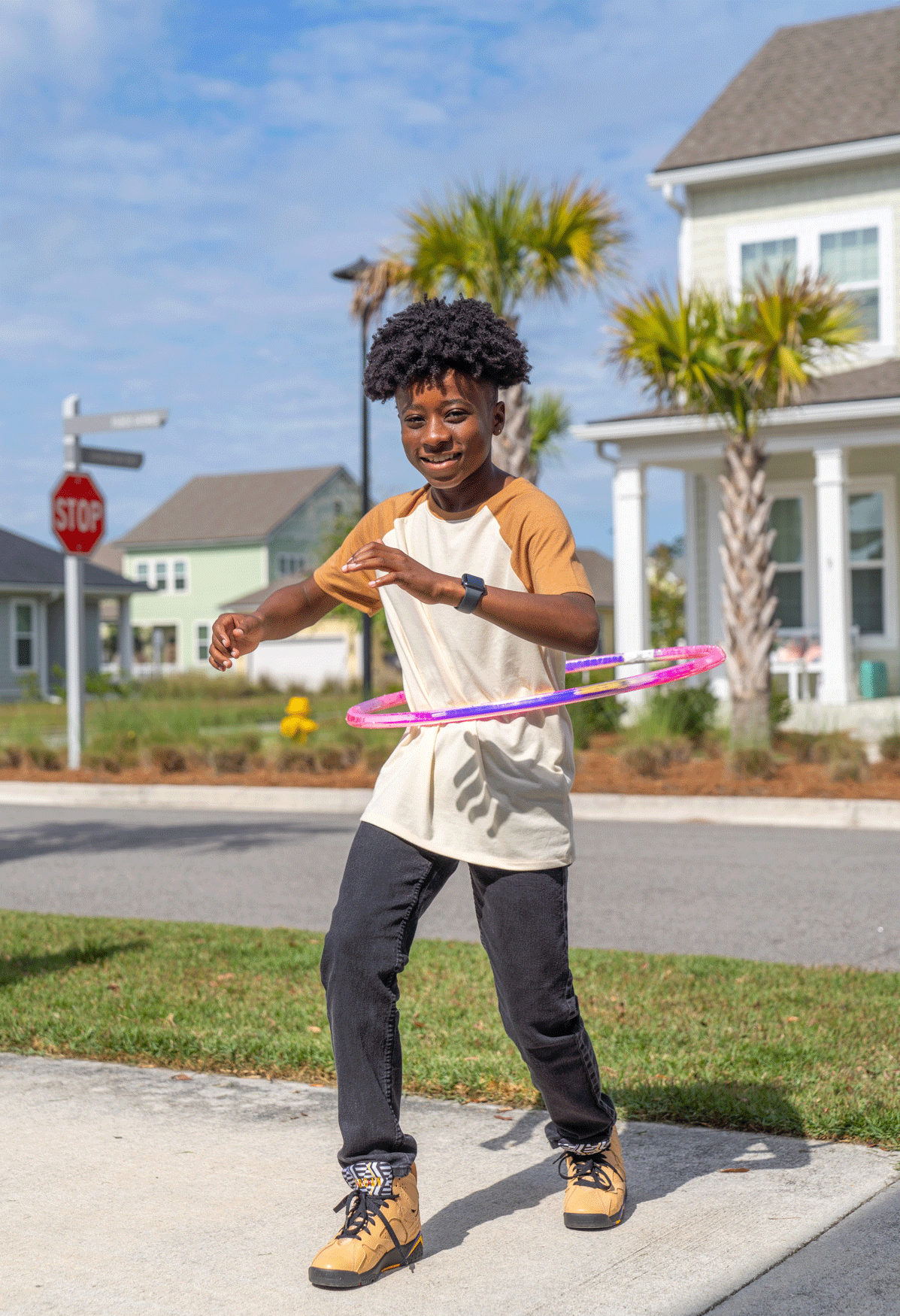 A young boy playing with a hula hoop on a sidewalk in a suburban neighborhood with palm trees, houses, and a stop sign in the background.