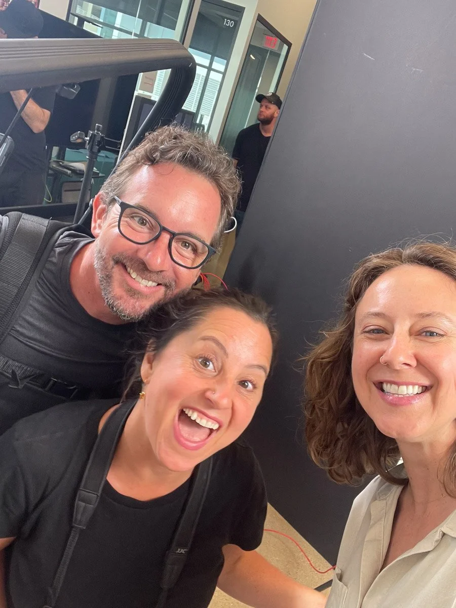 Three smiling adults taking a selfie together in an indoor setting, with a person and glass doors visible in the background.