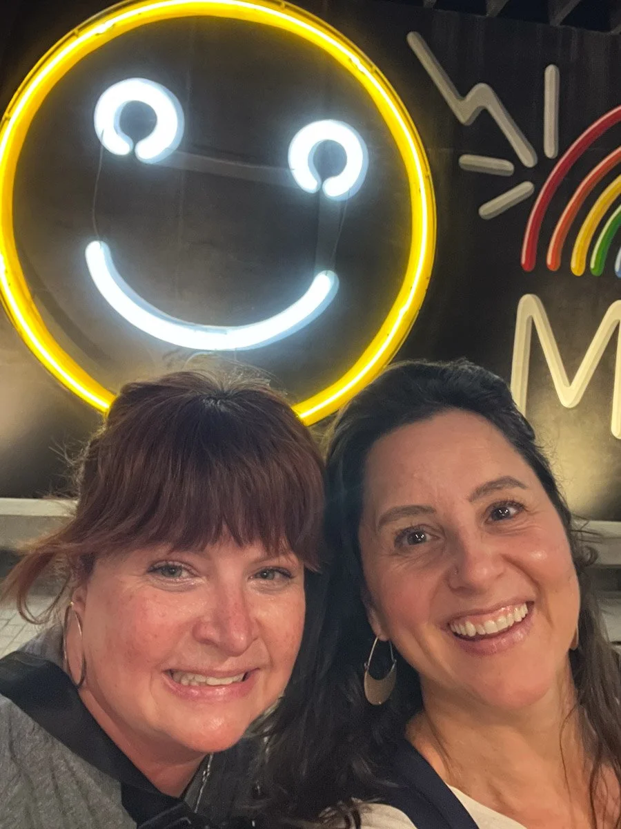 Two smiling women taking a selfie in front of a neon smiley face sign with a yellow outline. Part of a colorful rainbow mural is visible on the right side of the image.