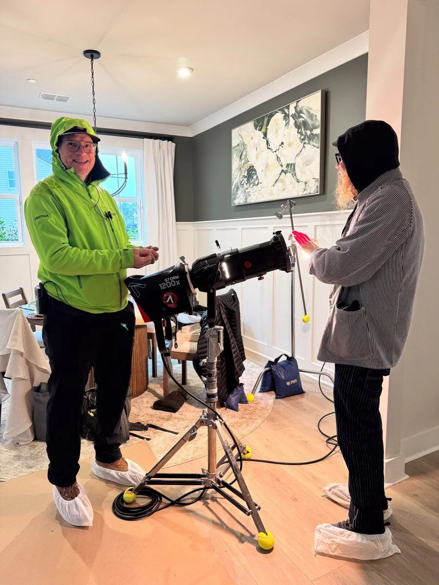 Two people in a living room with photography equipment. One person wears a bright green jacket and shoe covers, smiling at the camera; the other person wears a striped jacket, glasses, and pink gloves, holding his phone. There are lights, a camera, a